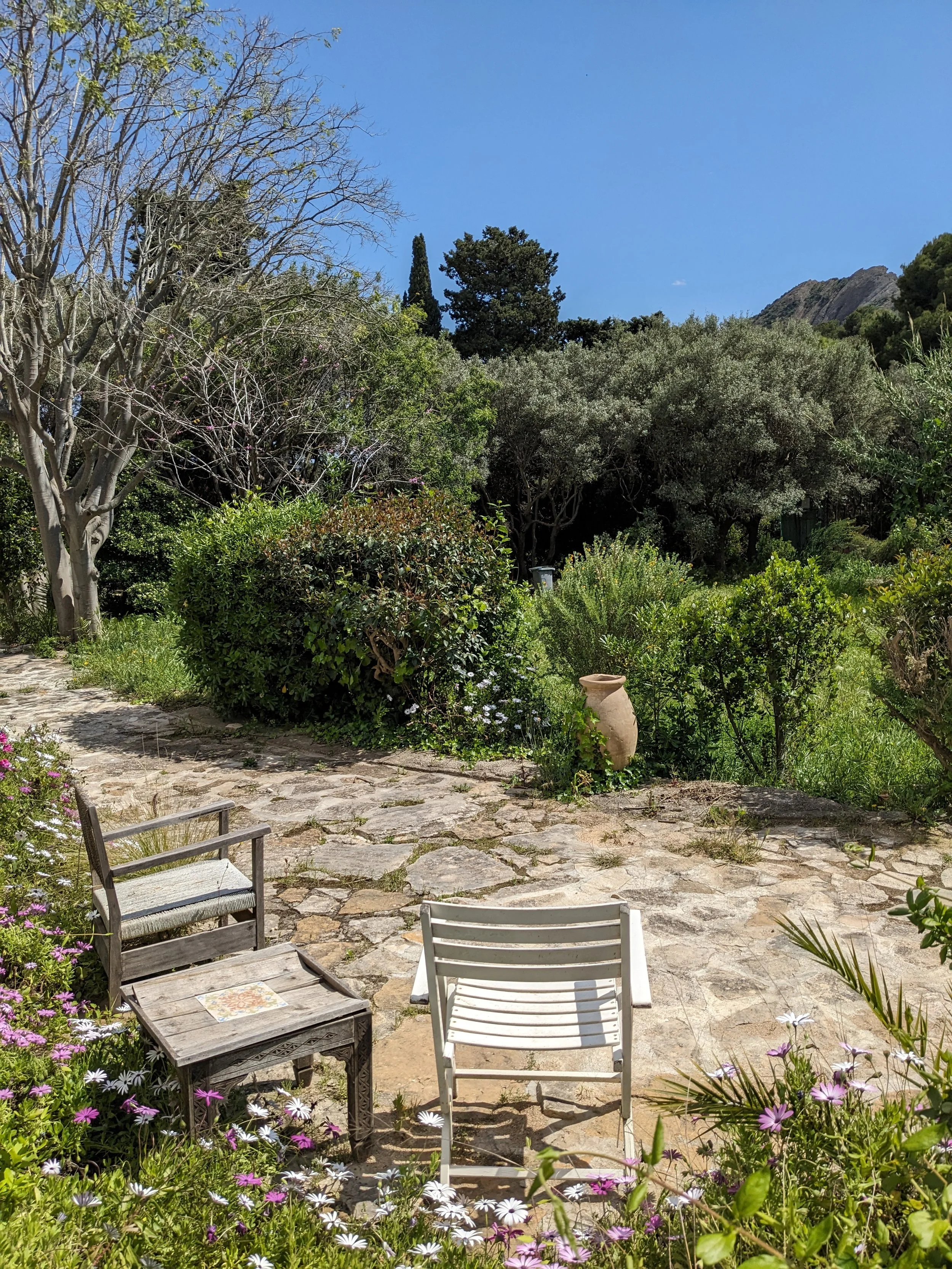 Ein Garten mit Steinterrasse, Stühlen, Blumen, Bäumen und einem Berg im Hintergrund bei schönem Wetter.
