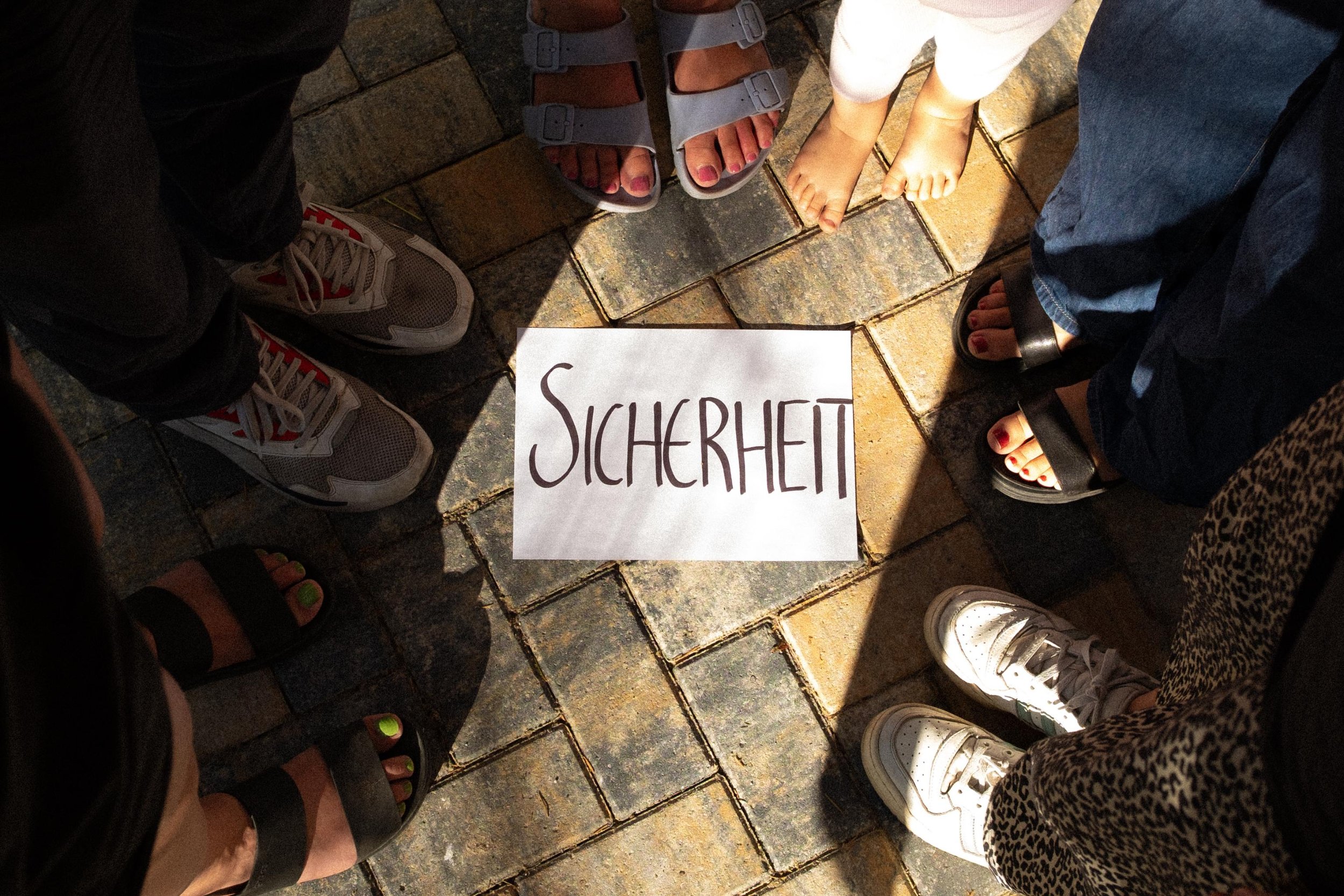 Füße mehrerer Menschen, die während eines Workshops im Kreis stehen. In der Mitte liegt ein handgeschriebener Zettel mit dem Wort 'SICHERHEIT'.