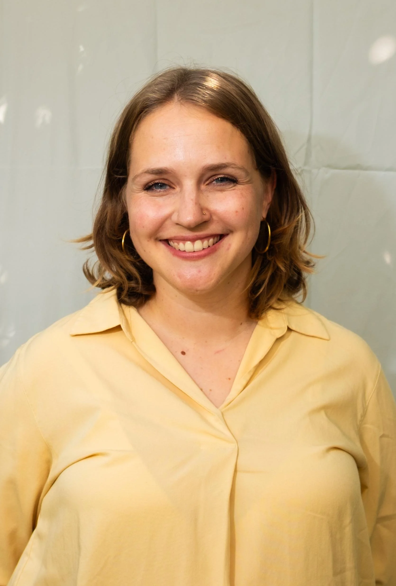 Porträt einer lachenden jungen Frau mit lockigem, schulterlangem Haar, trägt goldene Creolen und eine gelbe Bluse vor einem neutralen Hintergrund.