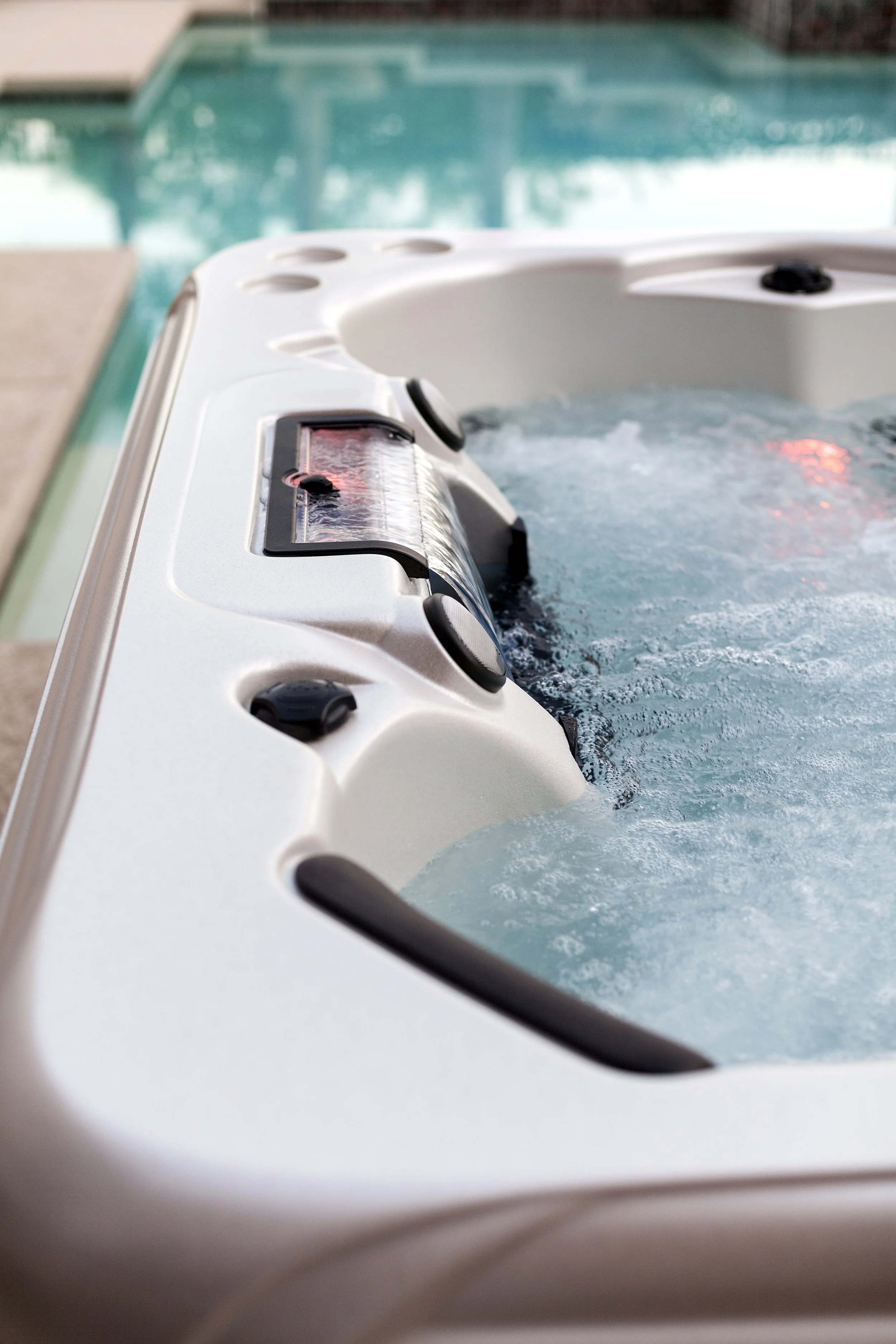Close-up of a white hot tub with bubbling water, showing control panel and jets, overlooking a pool and outdoor area.