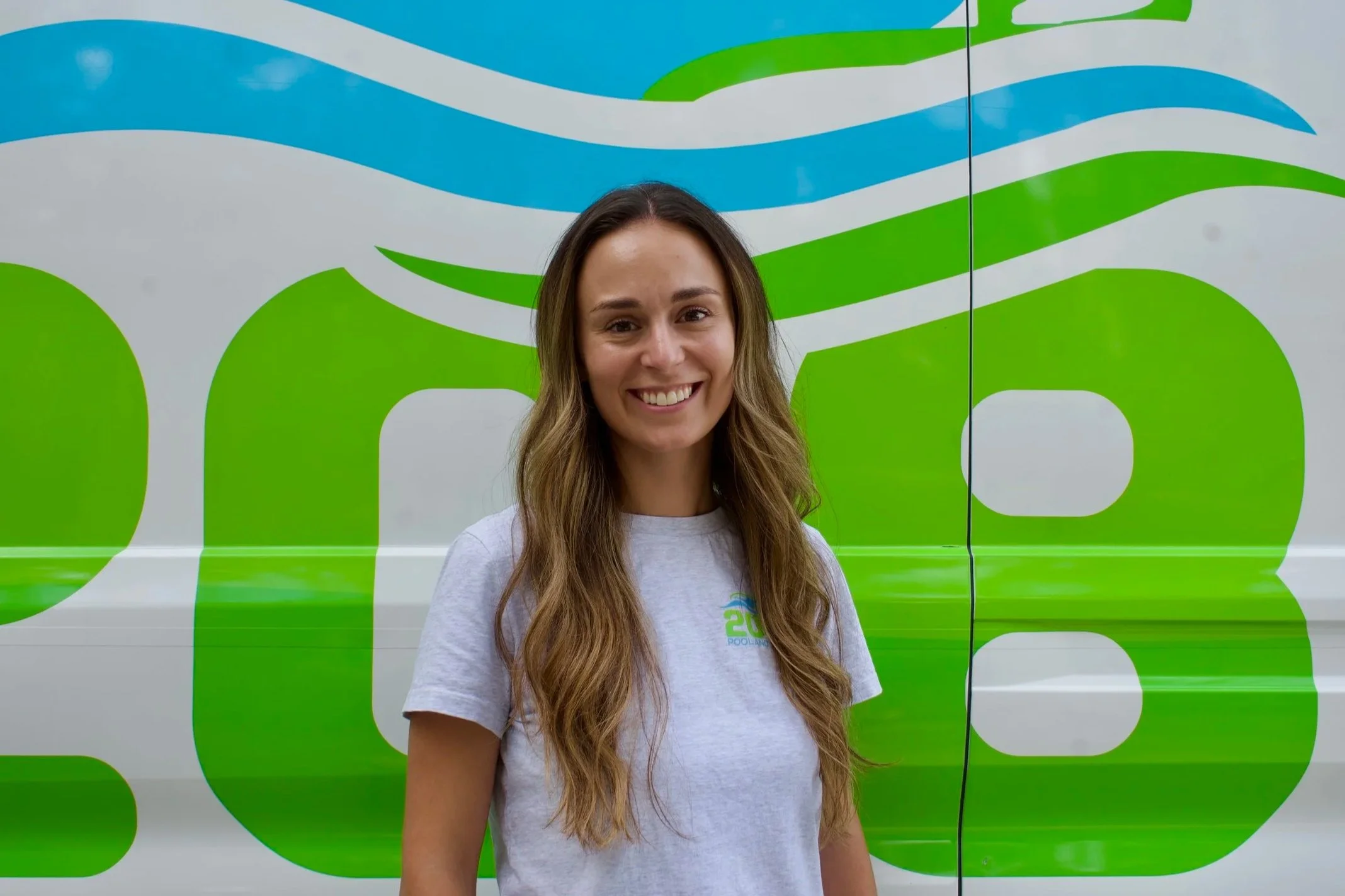 Smiling woman with long brown hair standing in front of a colorful abstract background painted on a wall or vehicle.