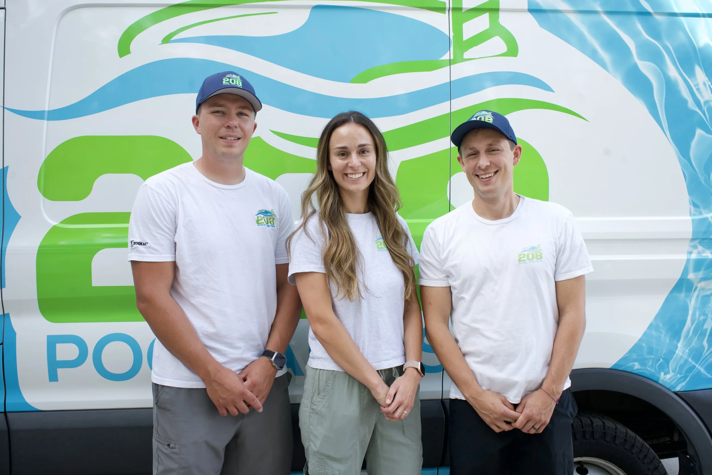 Three smiling people standing in front of a van with a colorful logo. The two men are wearing blue caps and white shirts with the same logo, while the woman is wearing a gray shirt with the same logo. The background features a blue and green design on the van.