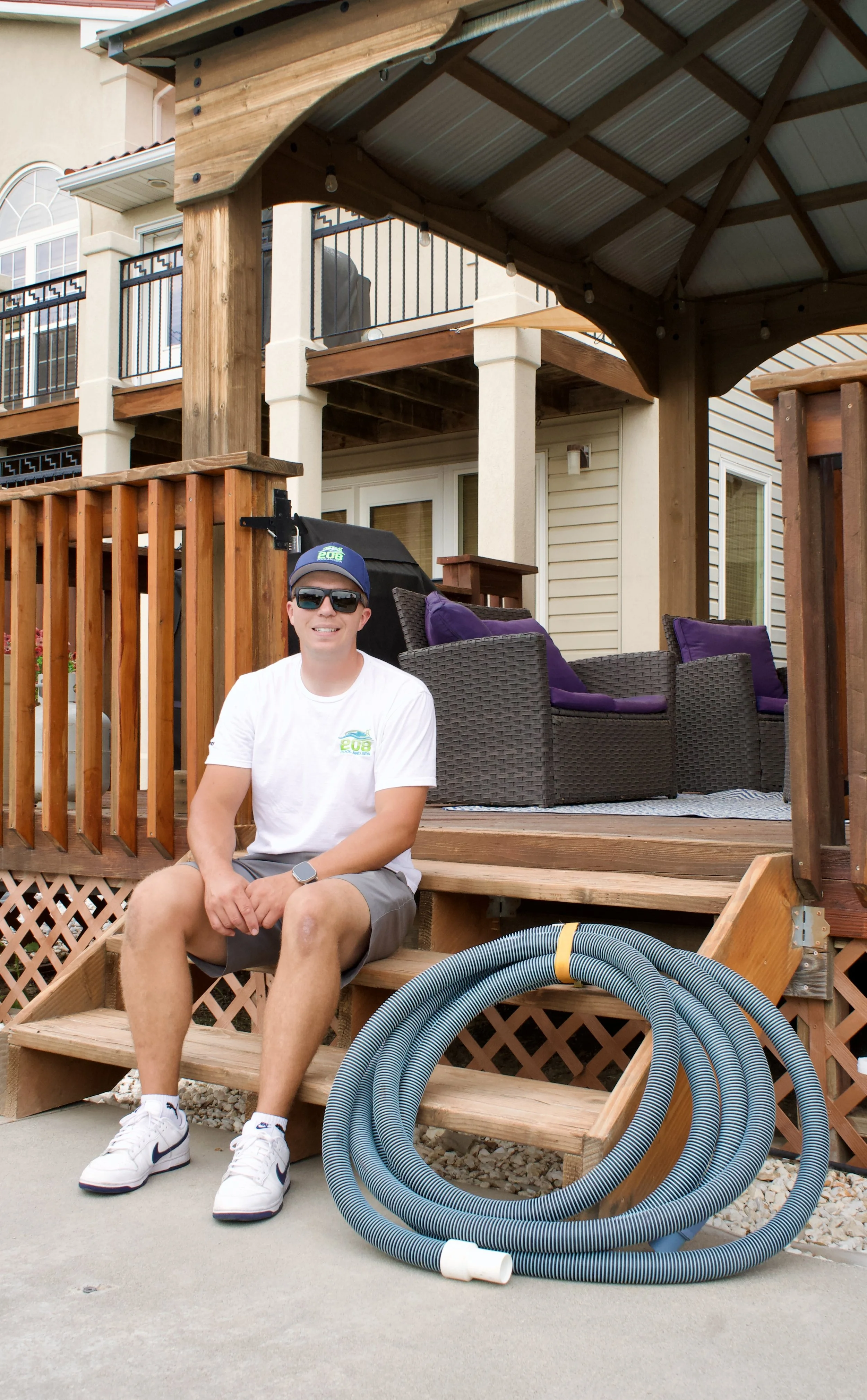 A man sitting on the steps of a wooden deck outside a house, smiling with sunglasses, wearing a white T-shirt, gray shorts, a baseball cap, and sneakers. The deck area has outdoor furniture with purple cushions, a barbecue grill, and garden hoses coiled on the ground.