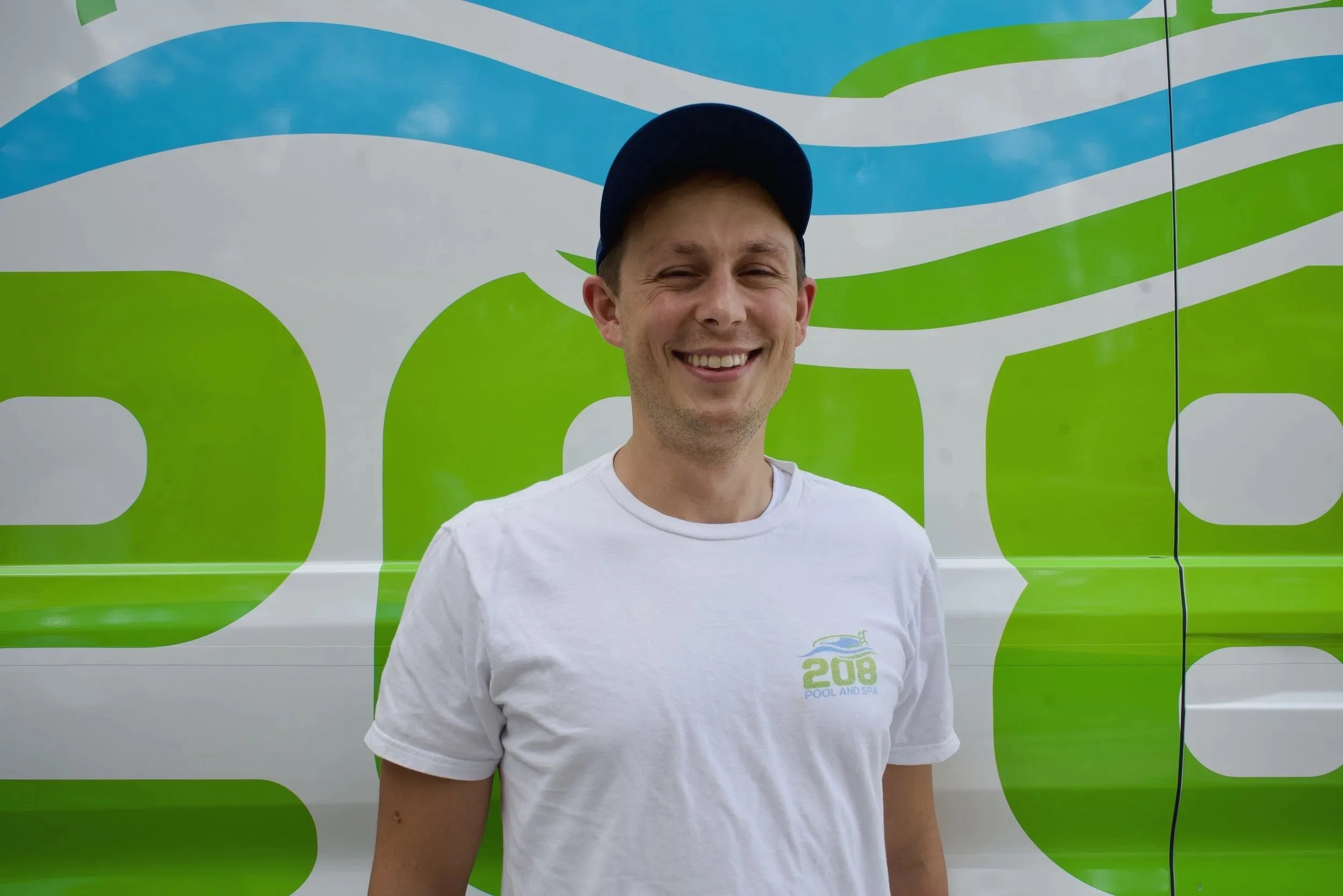 A young man smiling, wearing a white t-shirt with a logo and a blue cap, standing in front of a colorful wall with blue, white, and green wavy patterns.