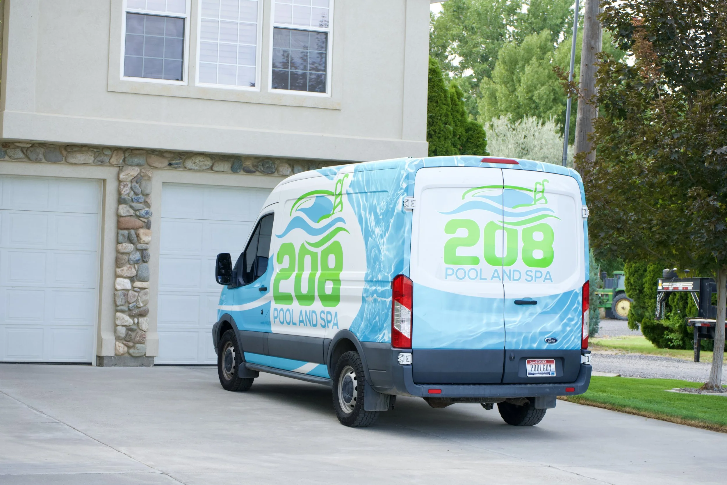 A white van with blue and green branding for 208 Pool and Spa parked in a driveway next to a residential house with a stone and stucco exterior, surrounded by trees and greenery.