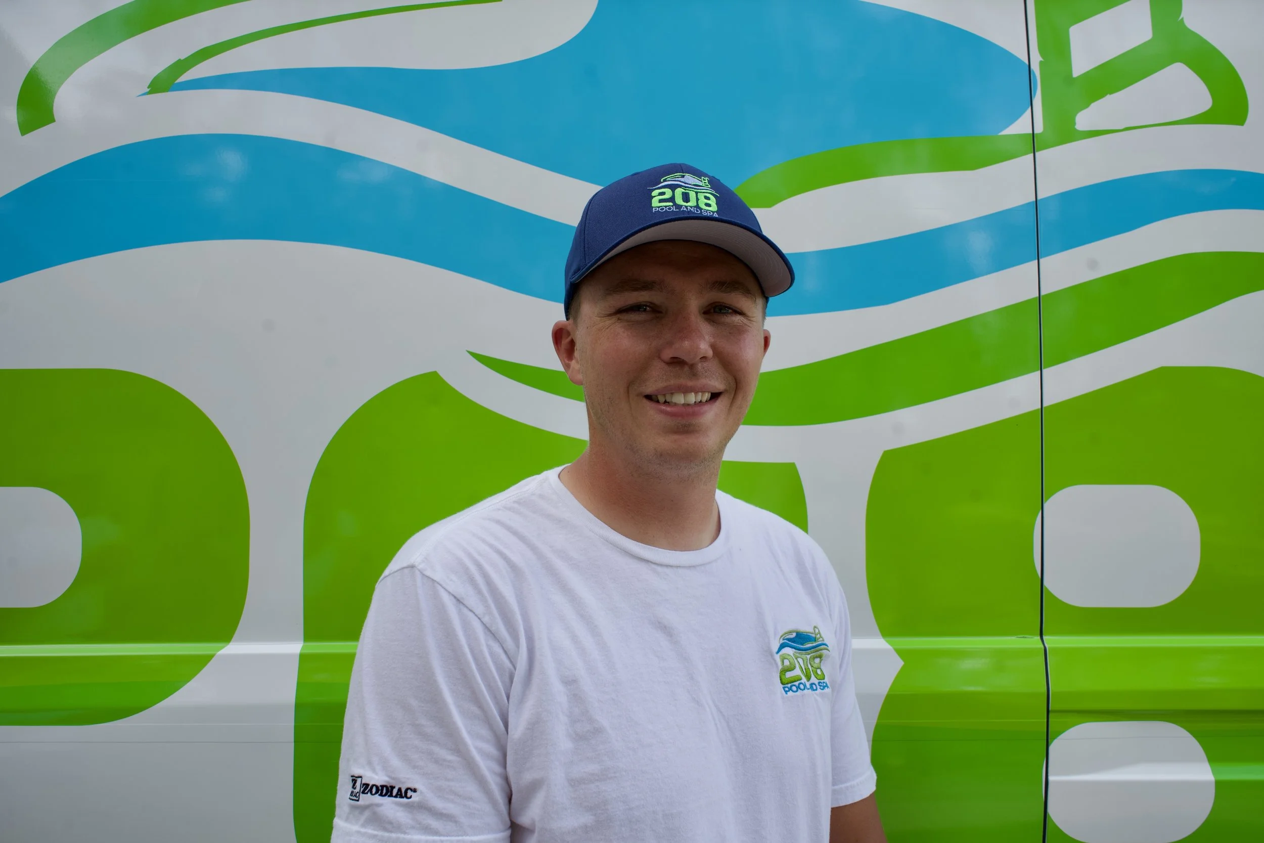 A smiling man wearing a navy blue cap and a white T-shirt in front of a blue, white, and green graphic background.