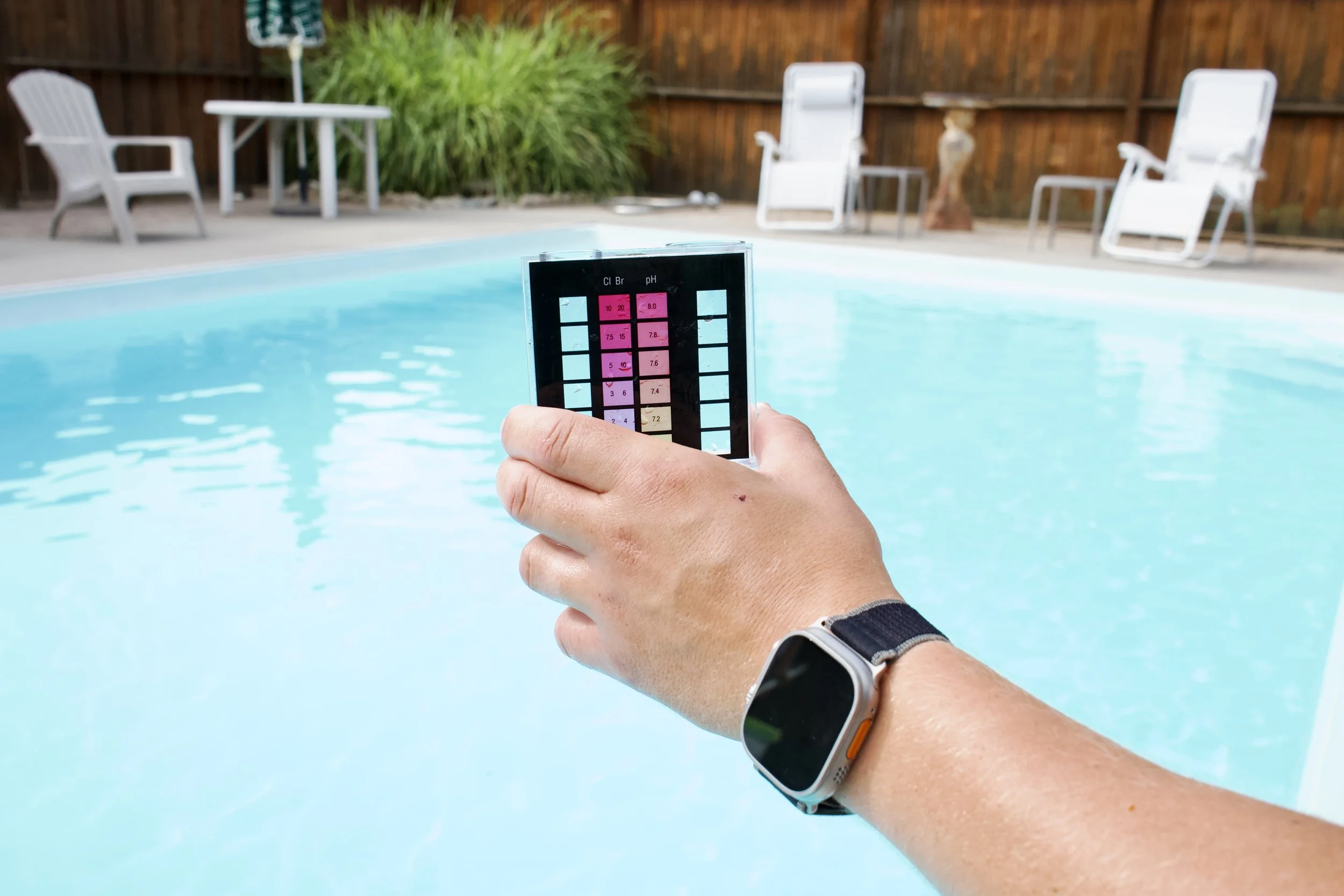 Person holding a pool test strip near a swimming pool, with poolside chairs and greenery in the background.