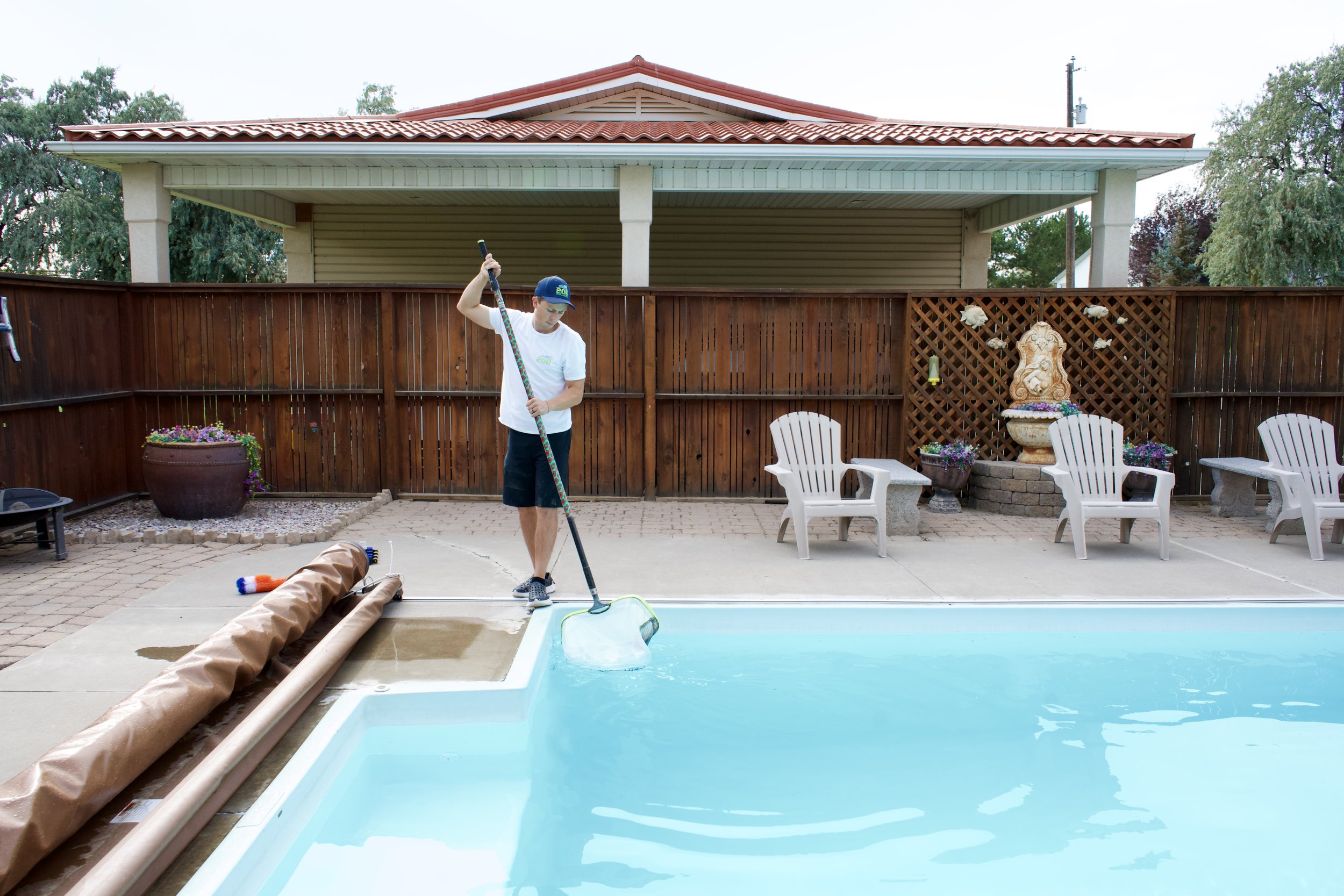 A person standing by an in-ground swimming pool, holding a pool skimmer, cleaning the pool. The backyard has a wooden fence, outdoor chairs, and decorative wall art.