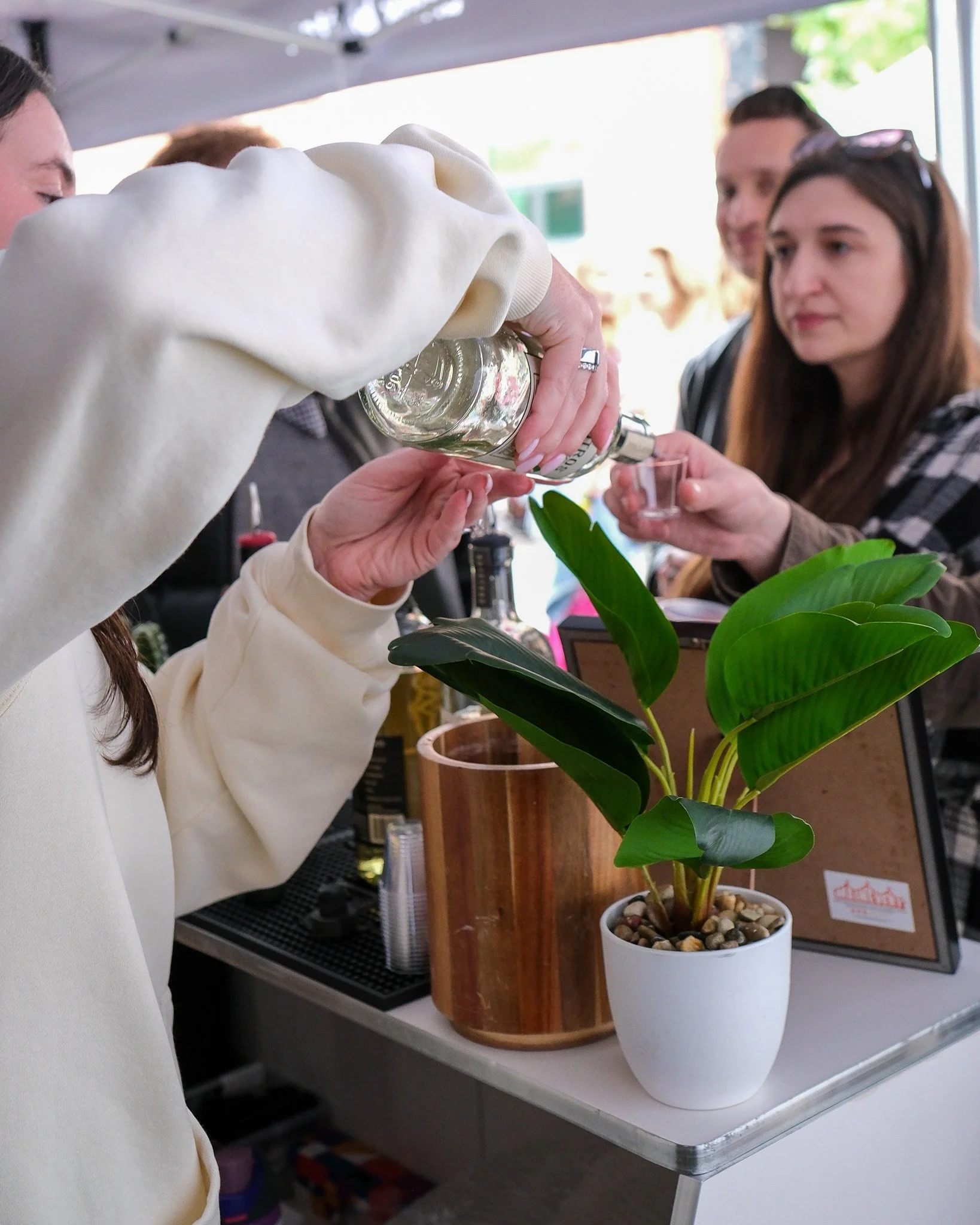People pouring clear liquid into small cups at an outdoor event with potted plants and a wooden podium.