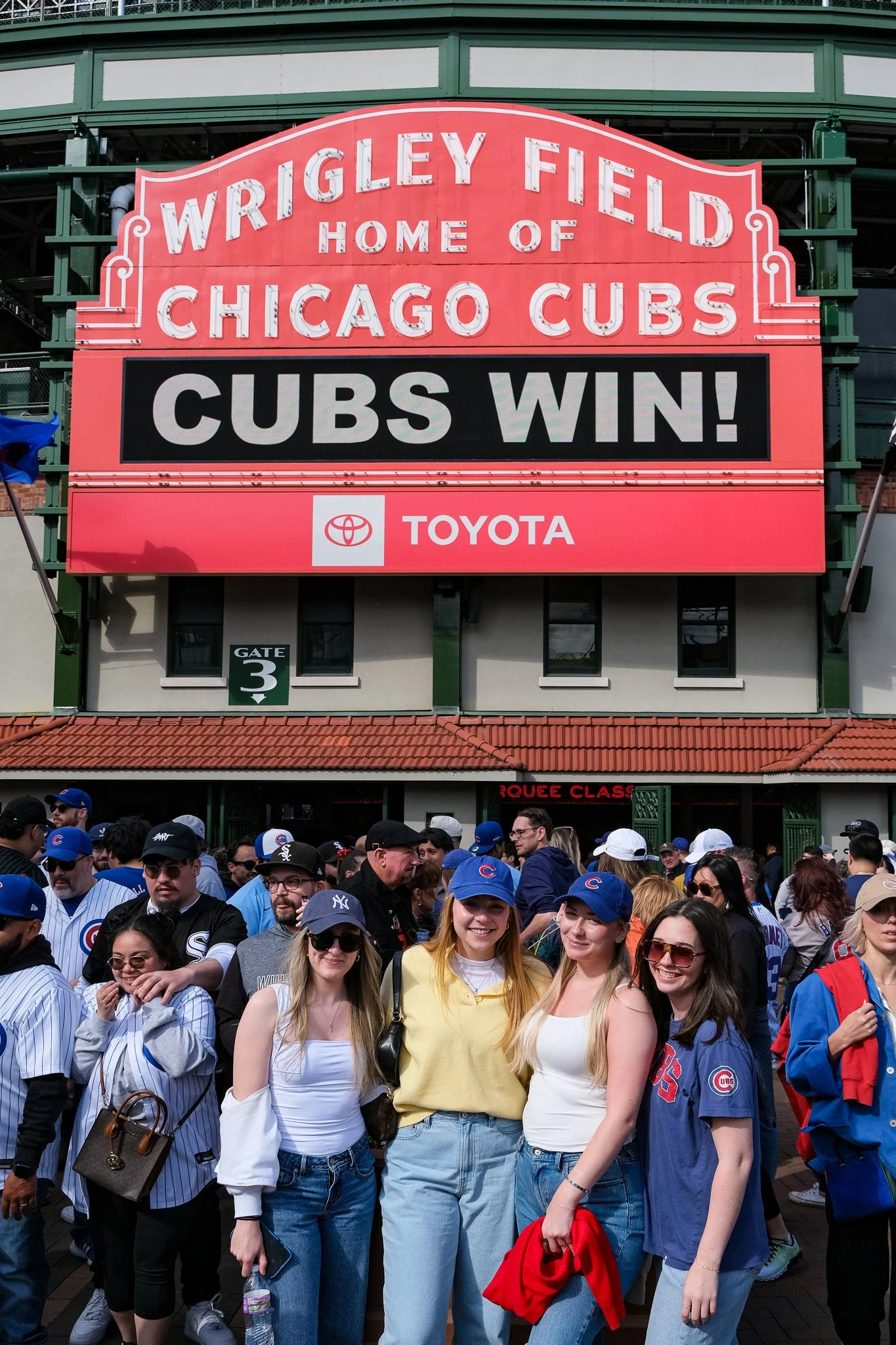 A crowd of people dressed in Chicago Cubs gear standing outside a stadium with a large sign that reads 'Wrigley Field Home of Chicago Cubs' and 'Cubs Win!'