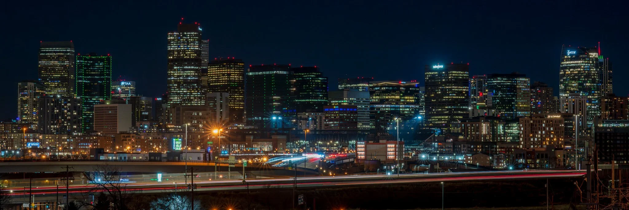 Nighttime city skyline with tall illuminated buildings, highway with light trails from moving vehicles, and a dark sky.