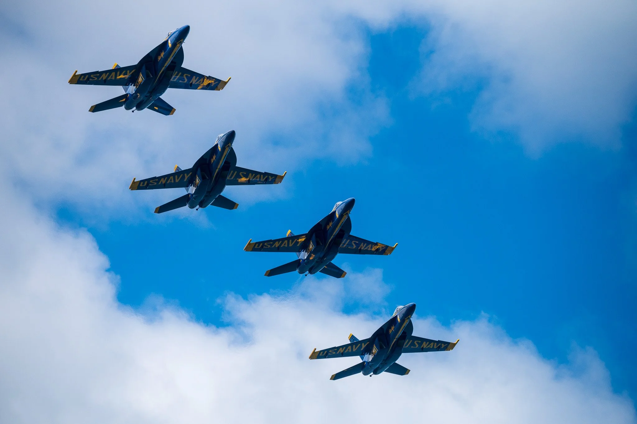 Four blue U.S. Navy fighter jets flying in close formation in a blue sky with some clouds.