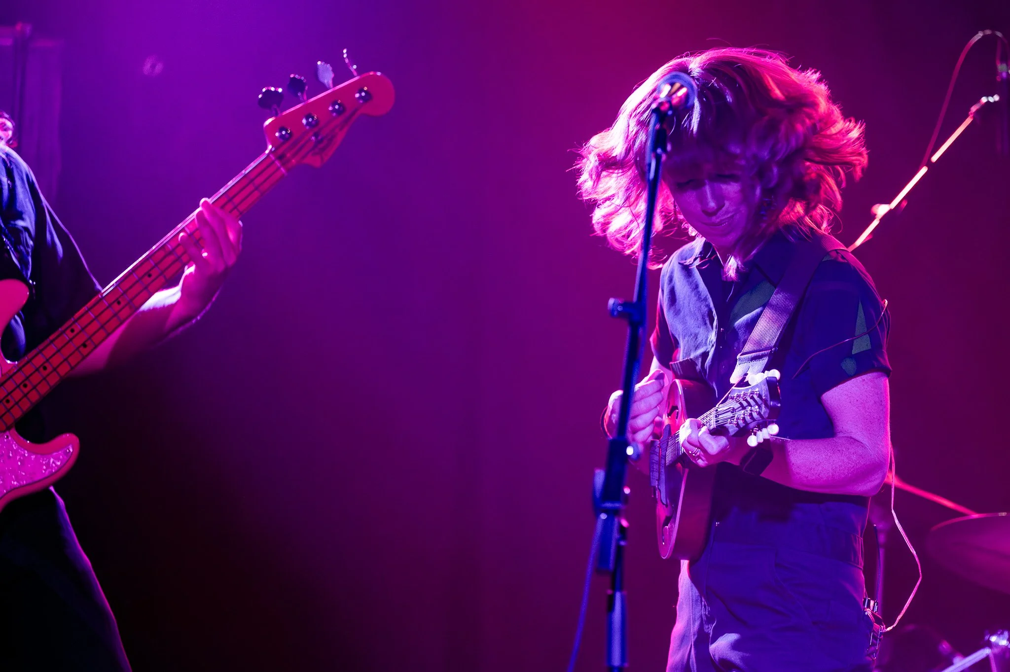 A woman with curly hair playing an acoustic guitar and singing into a microphone on stage with colorful purple and pink lighting.