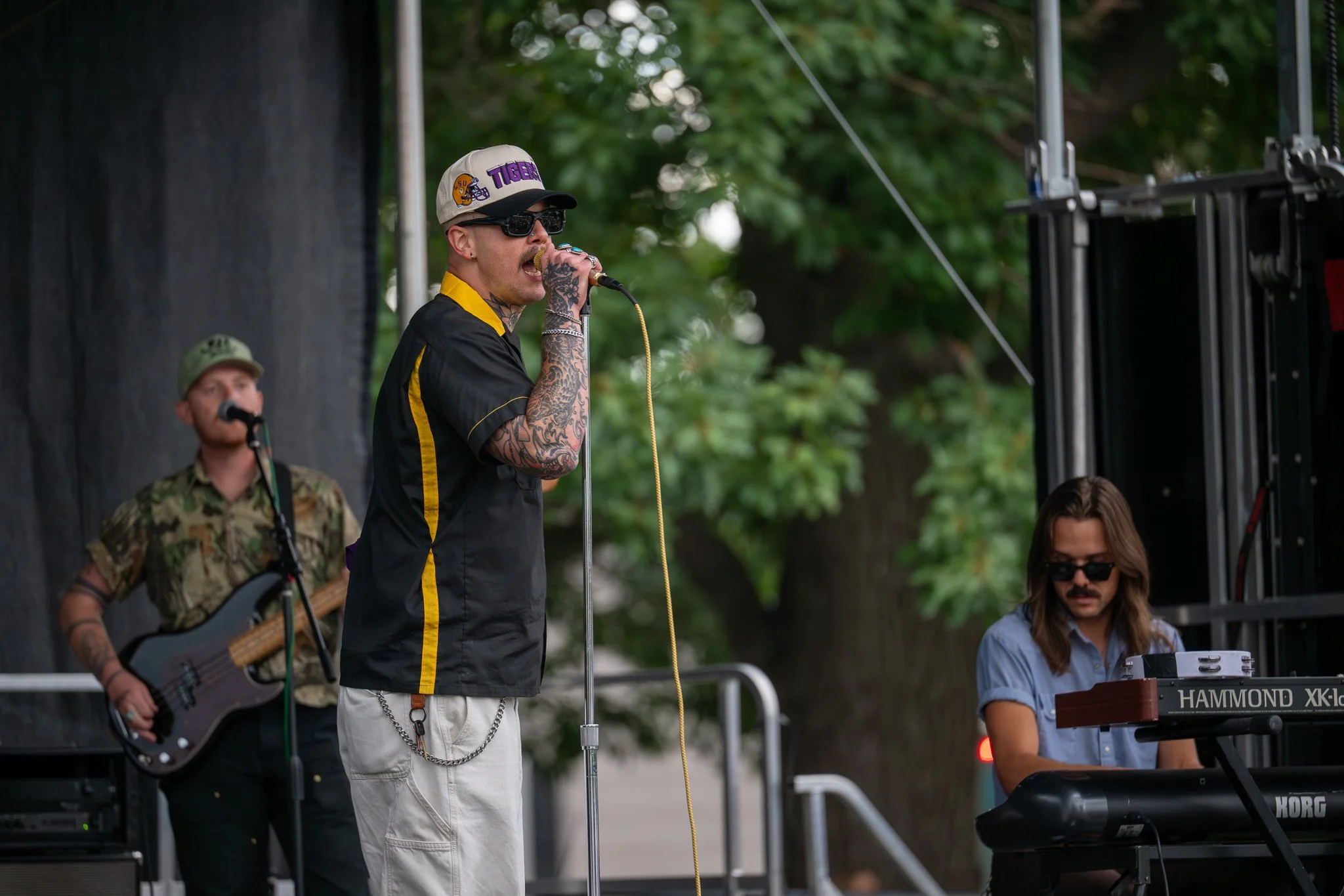A band performing outdoors with four members visible: a singer with tattoos, sunglasses, a baseball cap, and a microphone; a guitarist in camouflage shirt; a man with long hair and sunglasses playing keyboard; and a woman partially visible playing a 