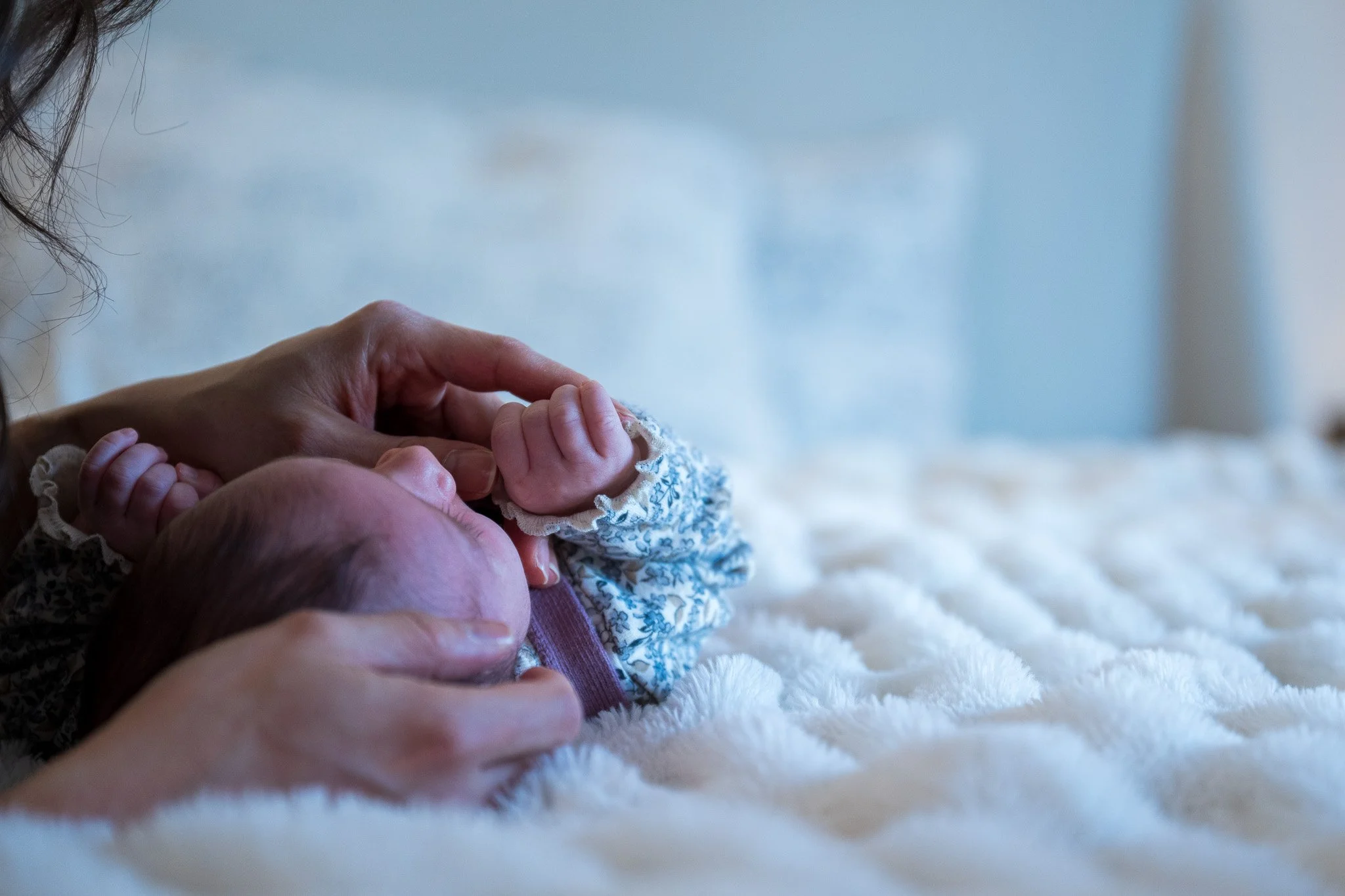 Close-up of a person holding a newborn baby's hand, both lying on a soft white surface, with the focus on their hands.