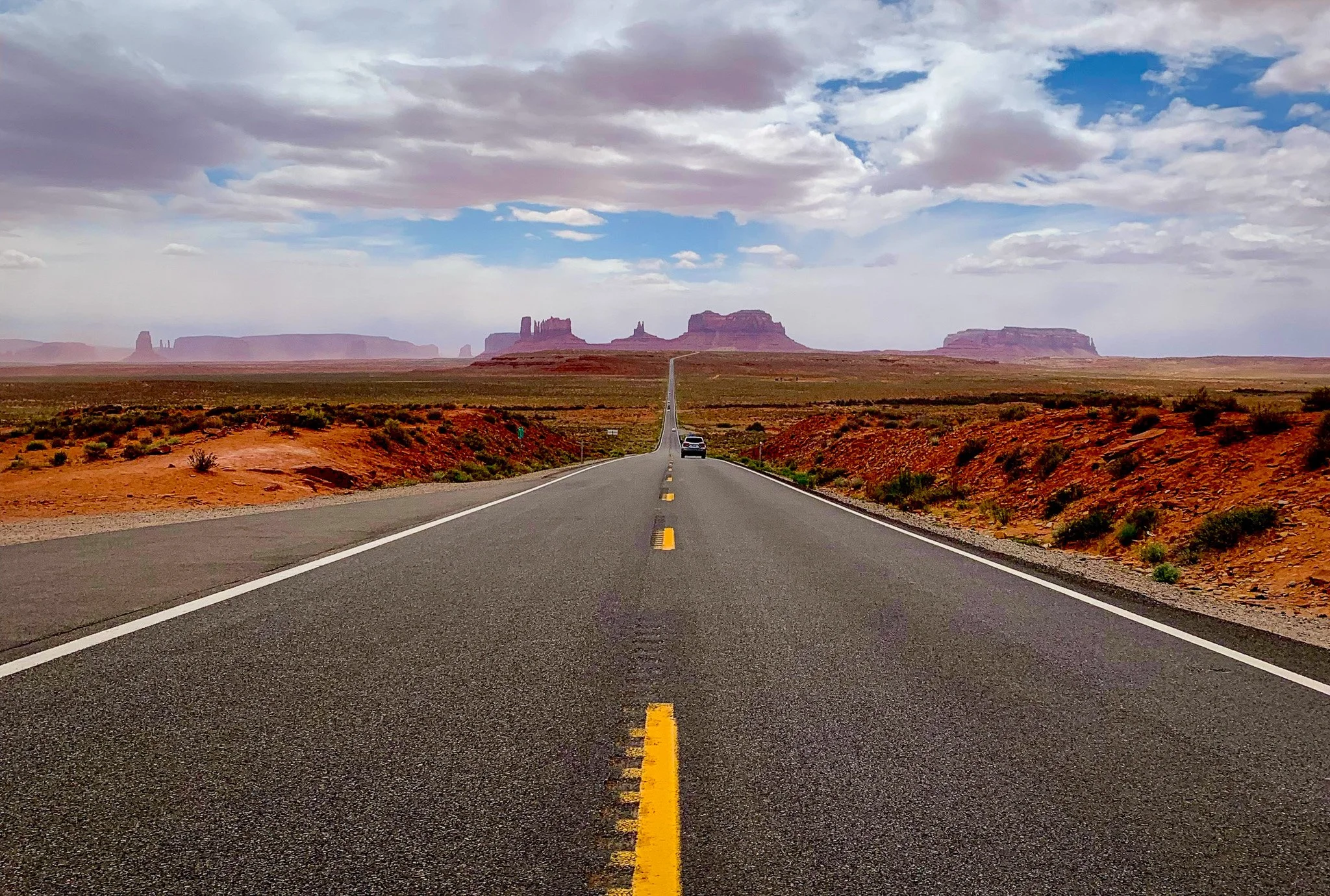 Long open road through desert landscape with iconic buttes and mesas in the distance, under partly cloudy sky.