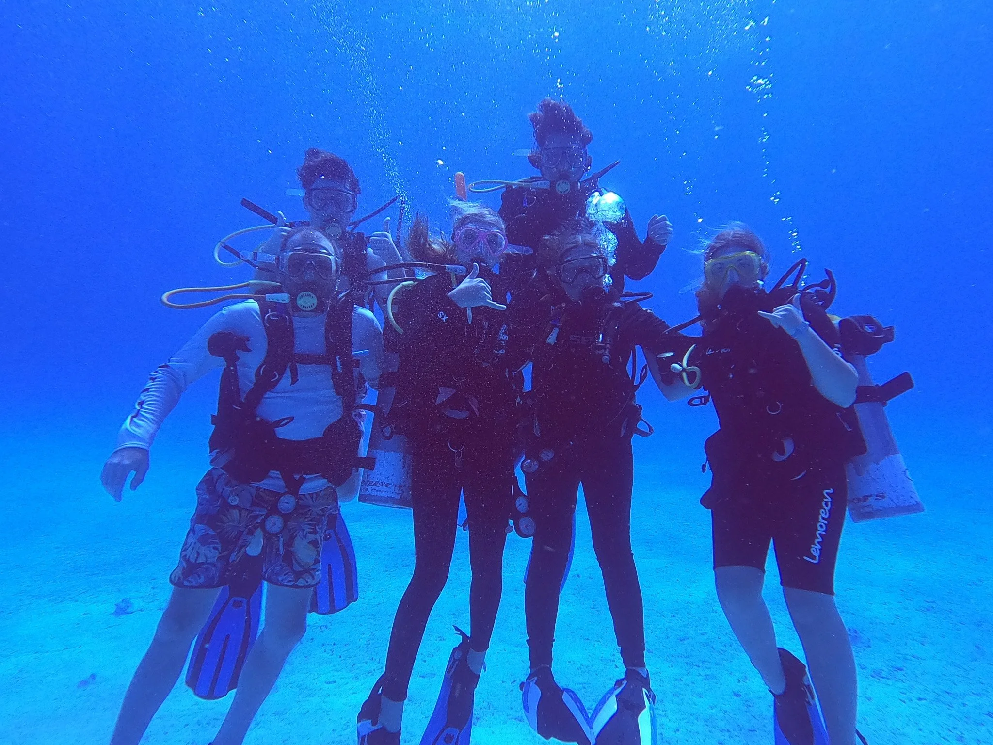 A group of five scuba divers underwater making the thumbs-up sign, surrounded by a blue sea environment.