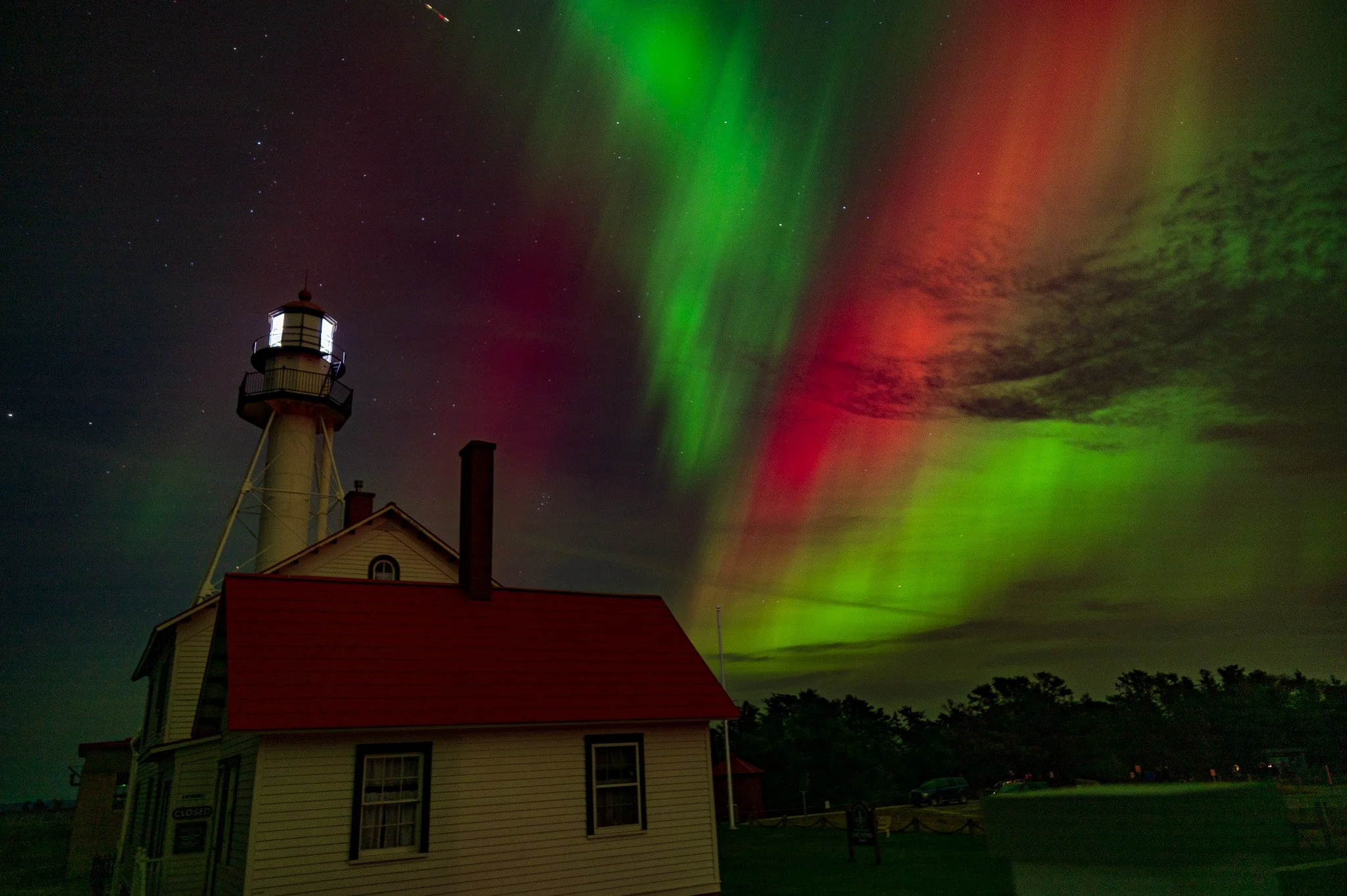 Night scene of a lighthouse with colorful northern lights in green and red in the sky, and a few stars visible.