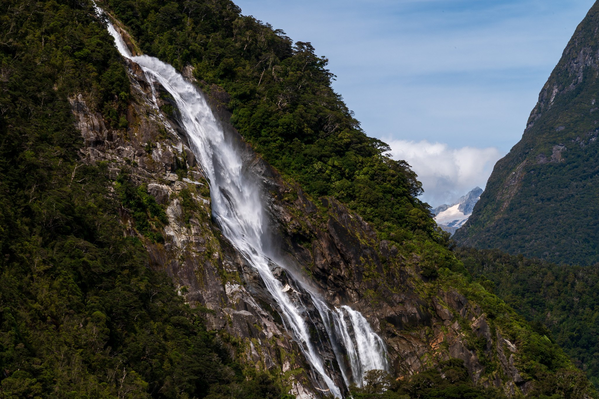 A waterfall flowing down a mountain surrounded by dense green forest with a partly cloudy sky in the background.