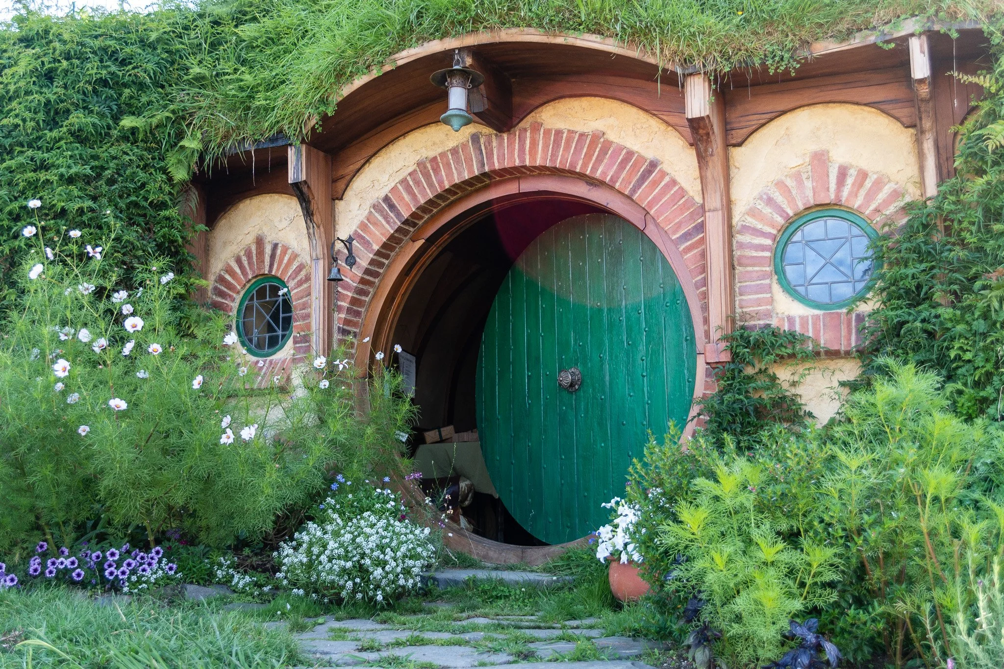 A Hobbit-style house entrance surrounded by lush green plants and colorful flowers, featuring a rounded green door with a decorative handle and circular windows with green frames.
