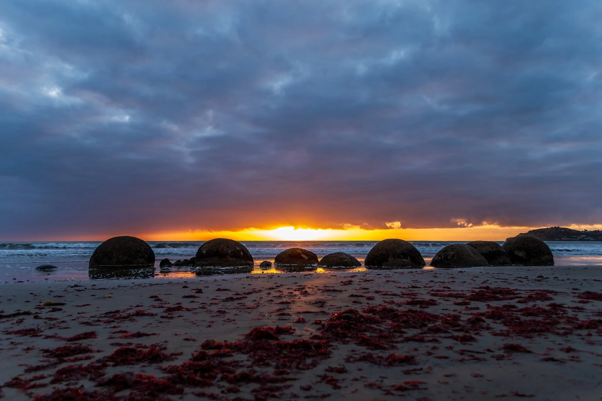 Beach scene during sunset with large rocks in the foreground, ocean waves, and a partly cloudy sky.