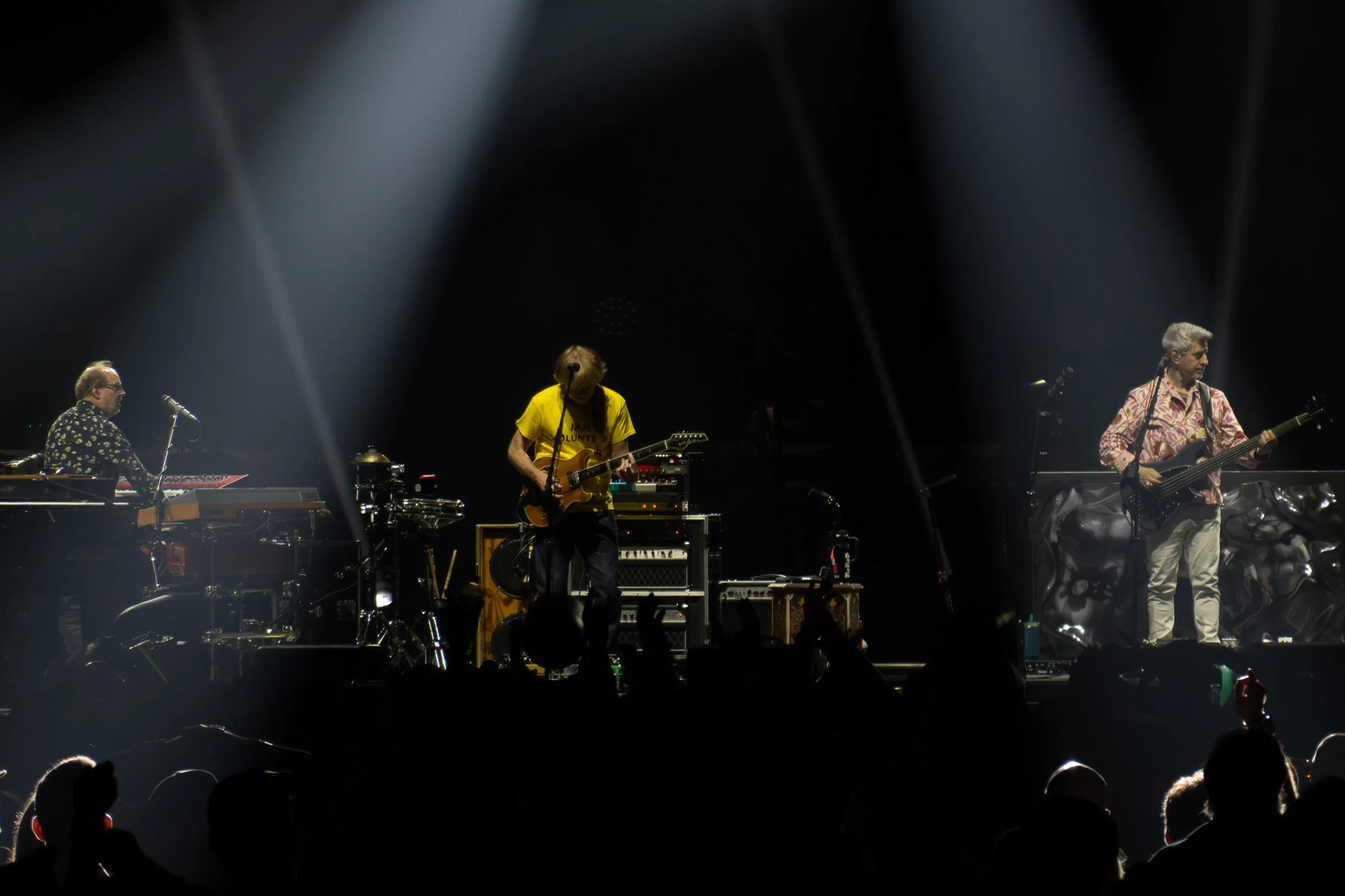 A band performing live on stage with multiple musicians playing guitars, keyboards, and drums, under dramatic lighting with hands and audience visible at the bottom.
