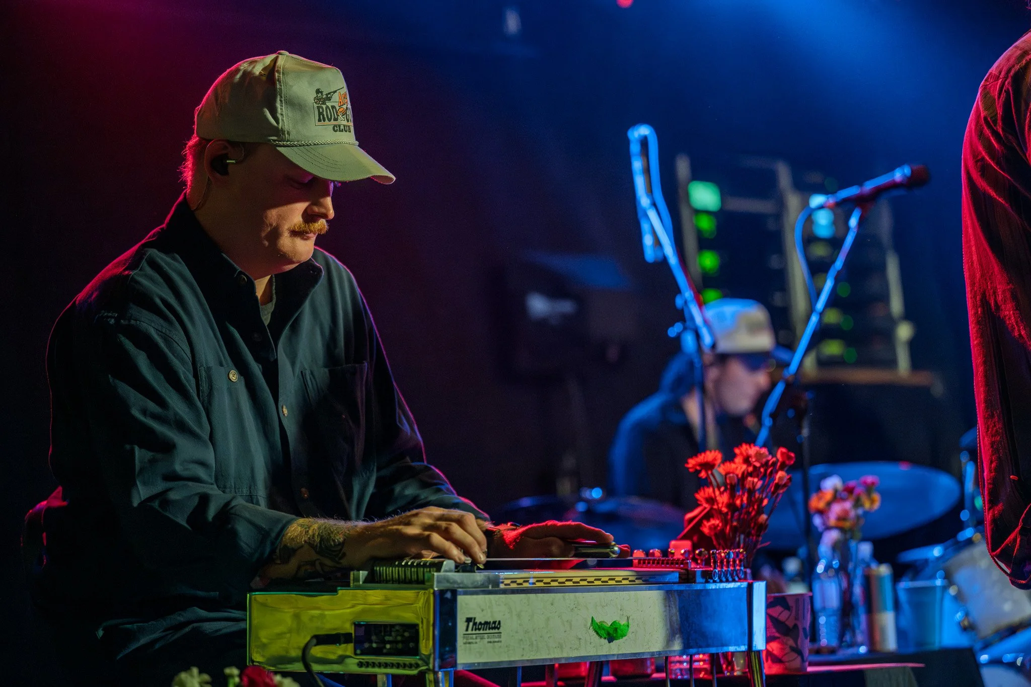 A man wearing a cap with a logo playing a keyboard on stage during a music performance, with colorful stage lighting and other band members in the background.