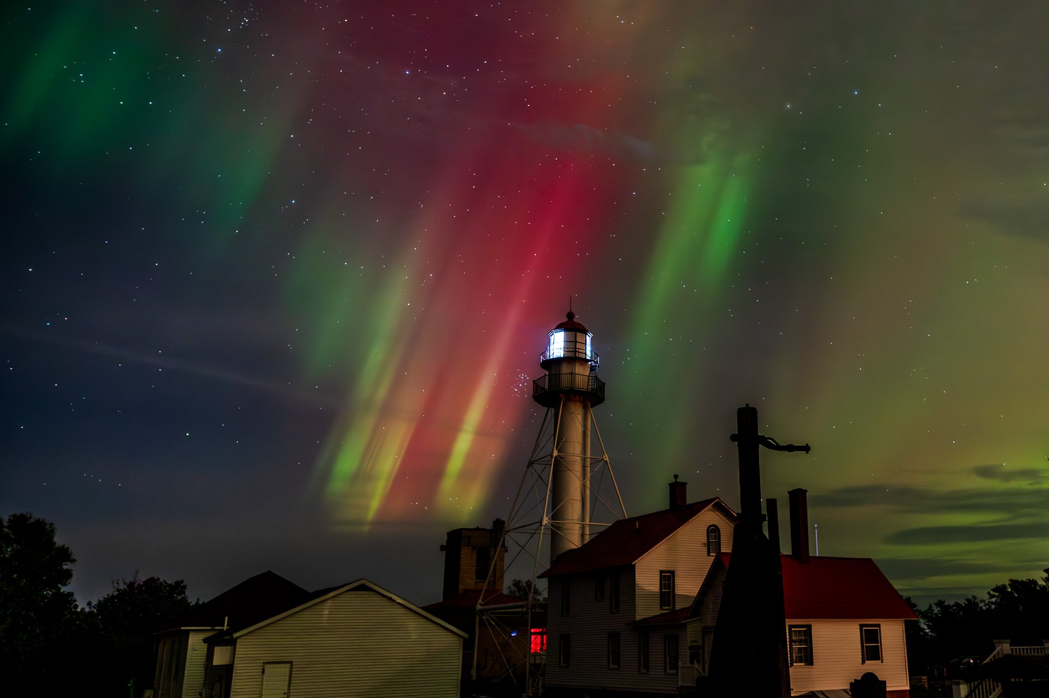 Night sky illuminated with colorful auroras above houses and a lighthouse.