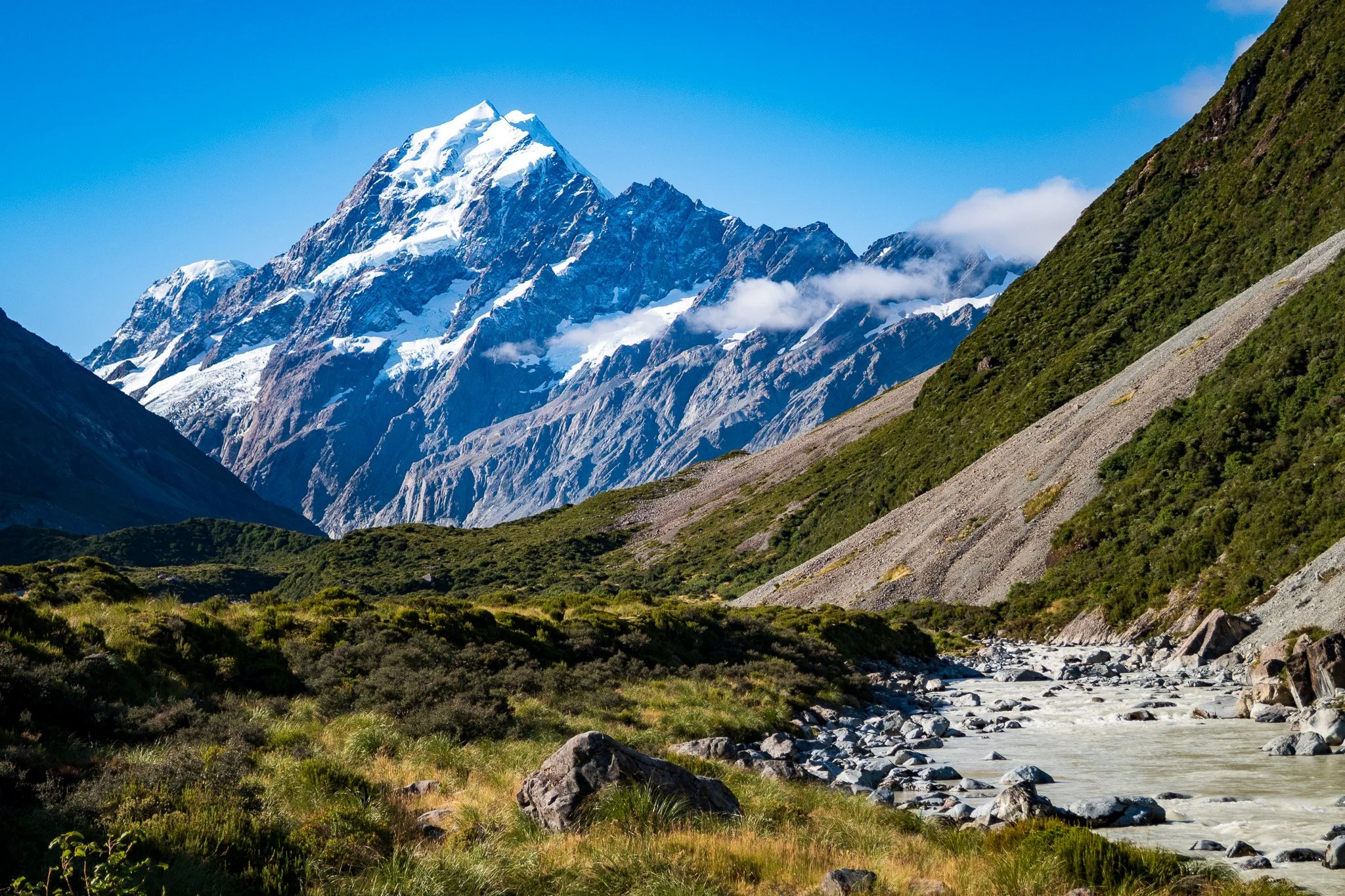 Tall snow-capped mountain peak rising above green hills and a rocky river valley.