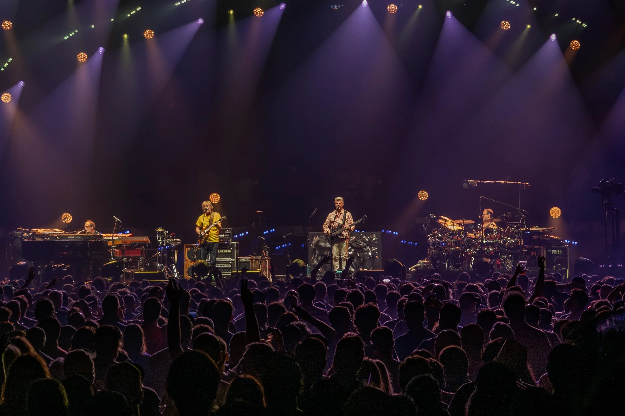 A live concert on stage with four band members performing. The stage is lit with colorful lights, and the audience is visible in the foreground with many hands raised.