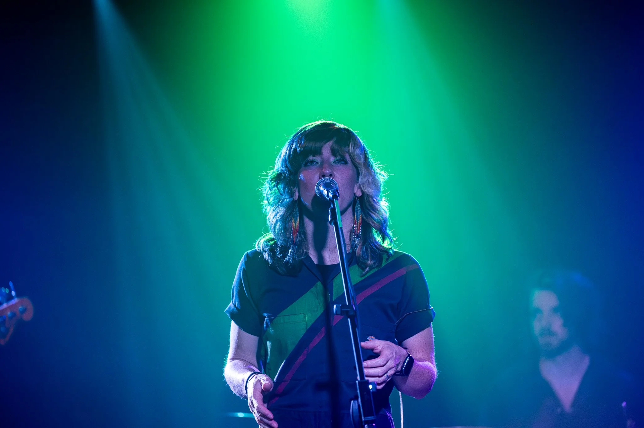 A woman with wavy, shoulder-length hair singing into a microphone on stage, illuminated by colorful stage lights with shades of green and blue.