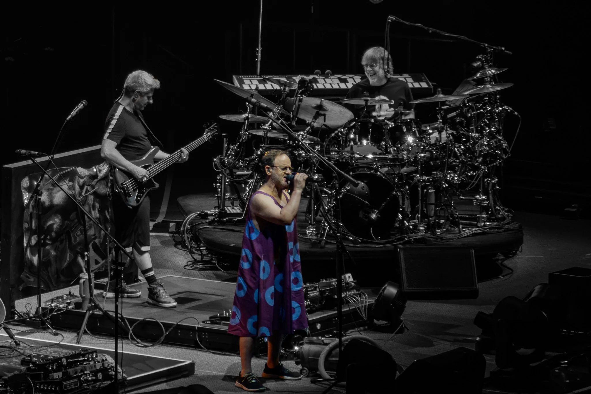 Three musicians on stage, one singing into a microphone and two playing instruments, with one visibly smiling. The stage has various electronic equipment and instruments.