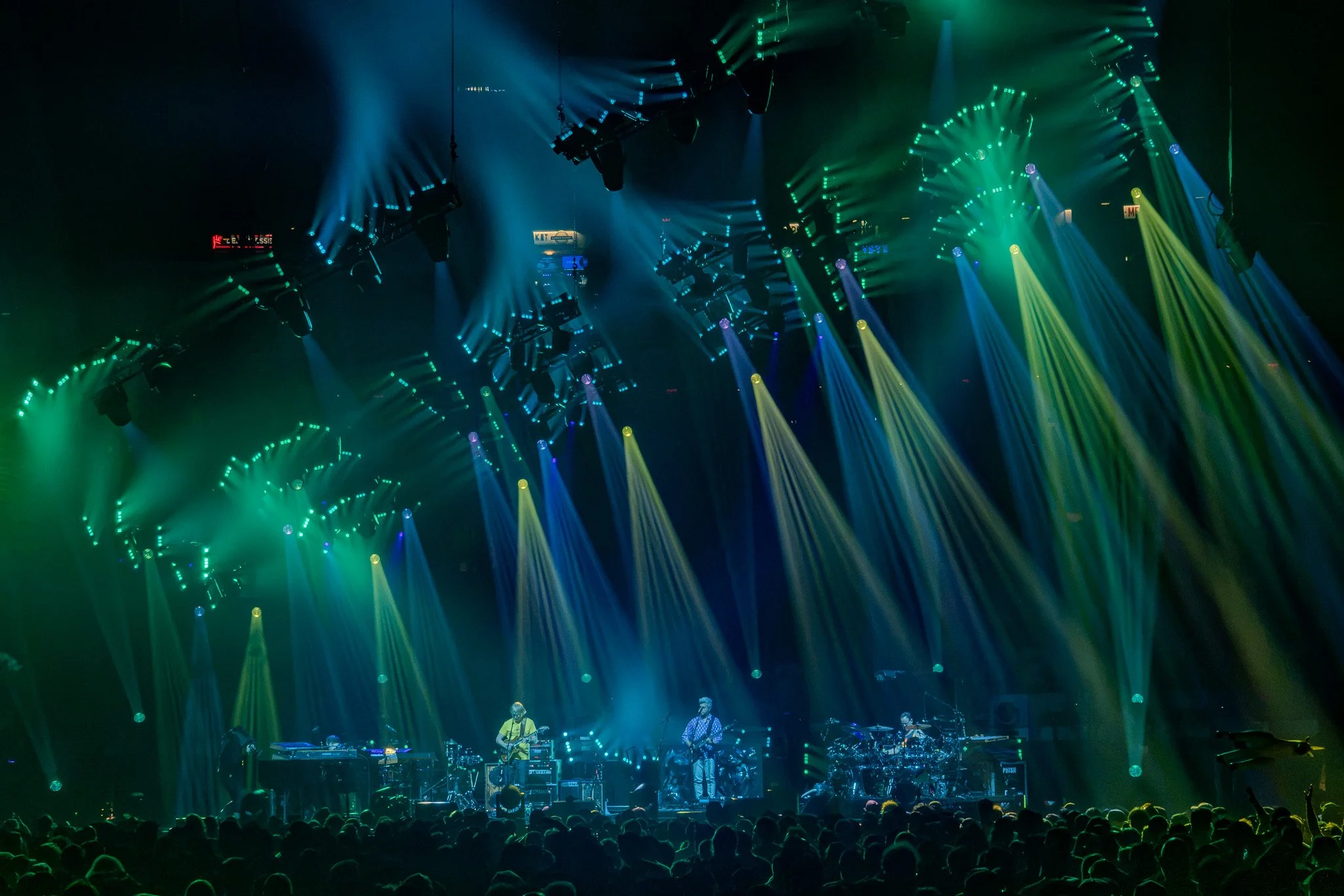 A concert stage illuminated with vibrant green, yellow, and blue lighting, with musicians performing and a large audience watching.