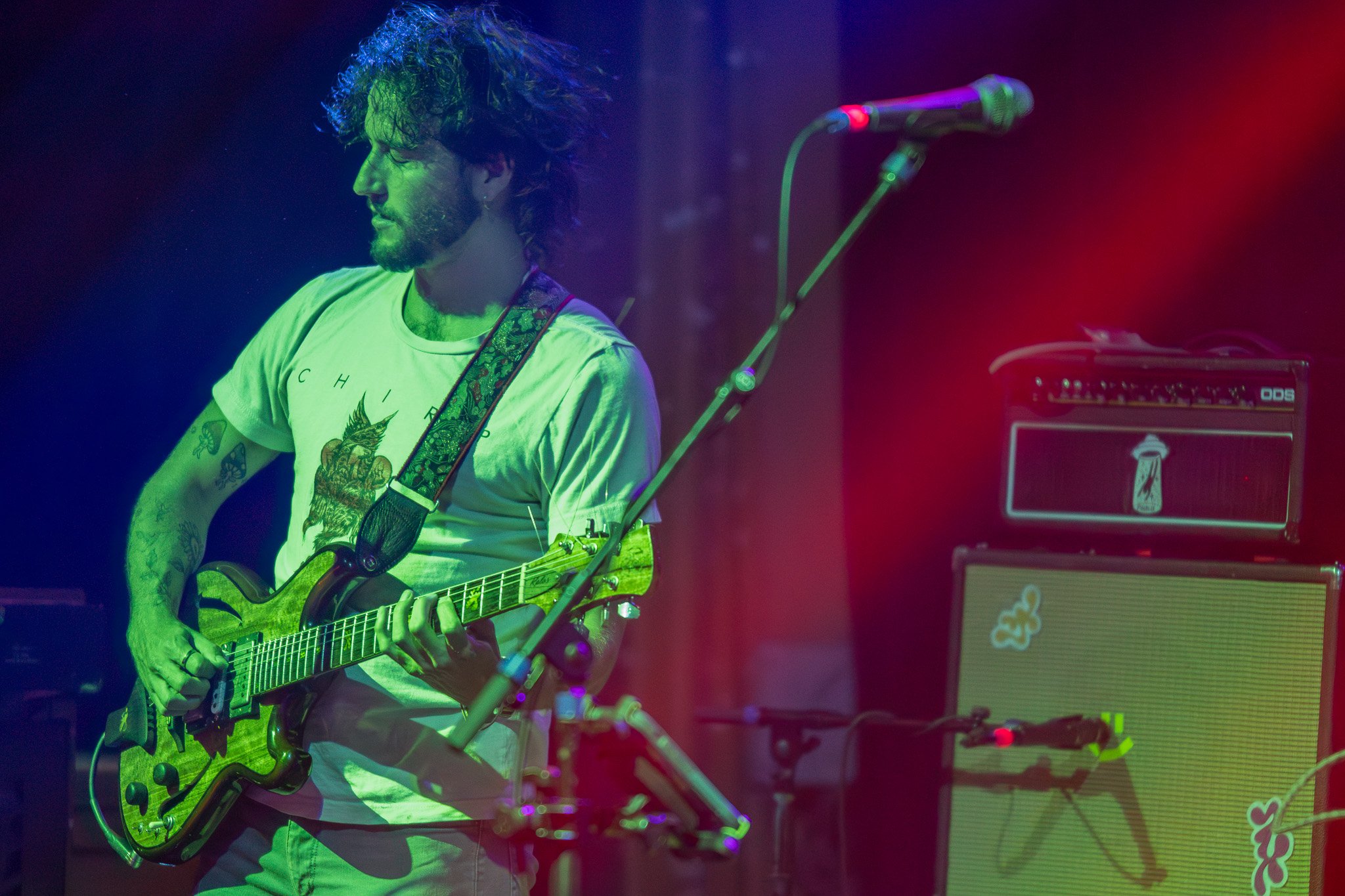 A man with curly hair playing an electric guitar on stage, illuminated by colorful stage lights, with a guitar amplifier in the background.