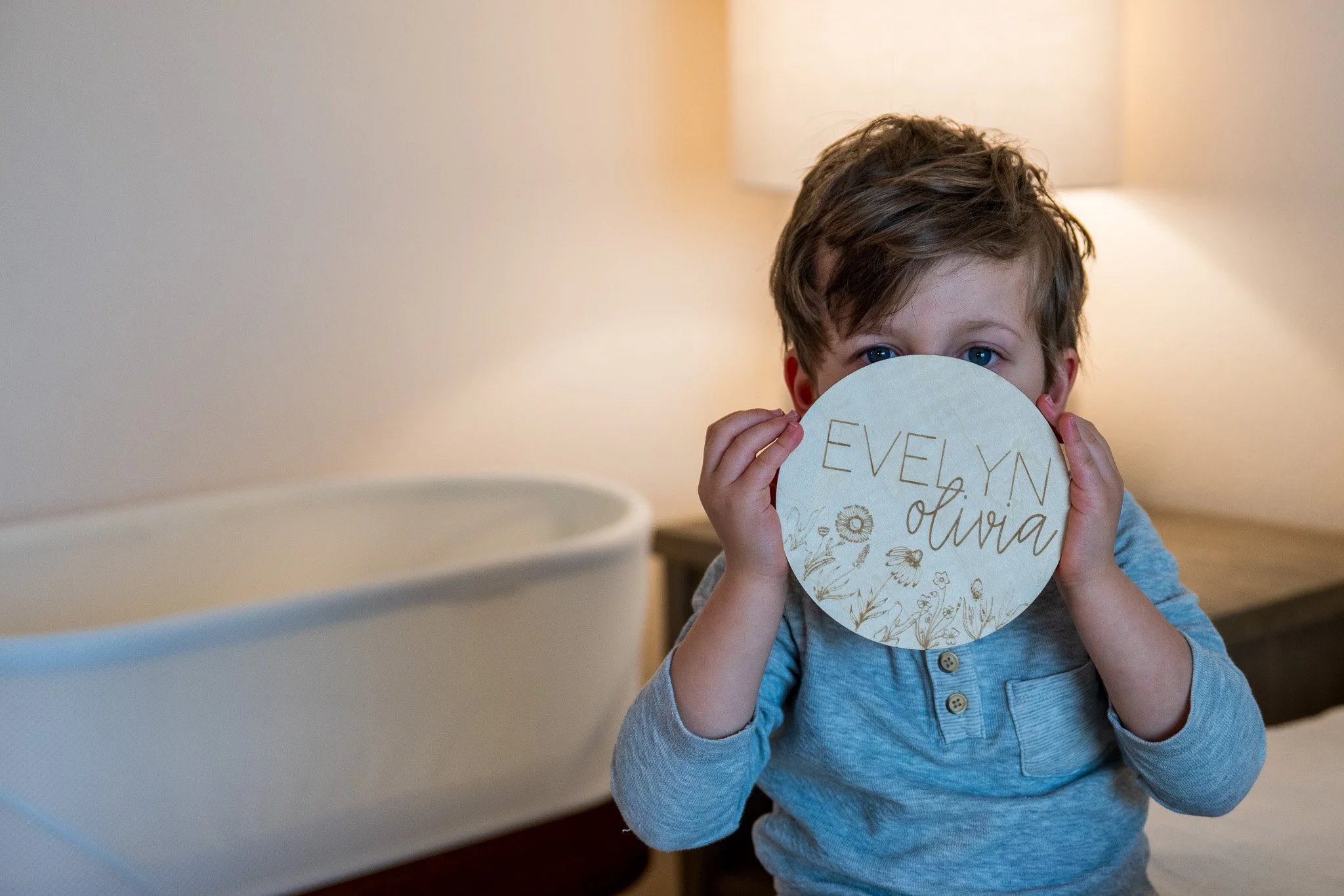 A young boy with curly brown hair and blue eyes holding a round card with floral designs and the names Evelyn and Olivia written on it, partially covering his face, sitting indoors near a lamp.