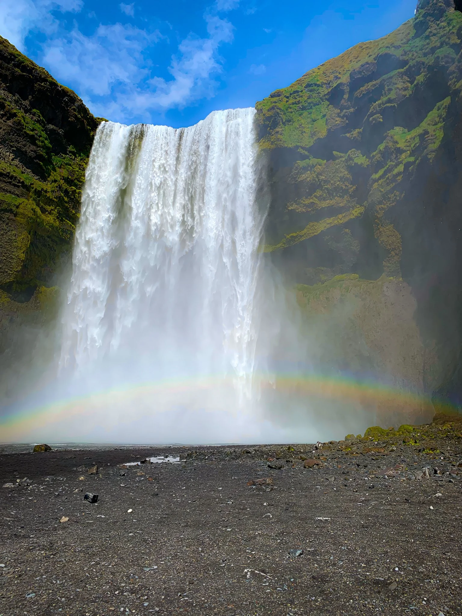 A large waterfall cascading down a green mountainside under a partly cloudy sky, with rainbows at the base over dark rocky ground.