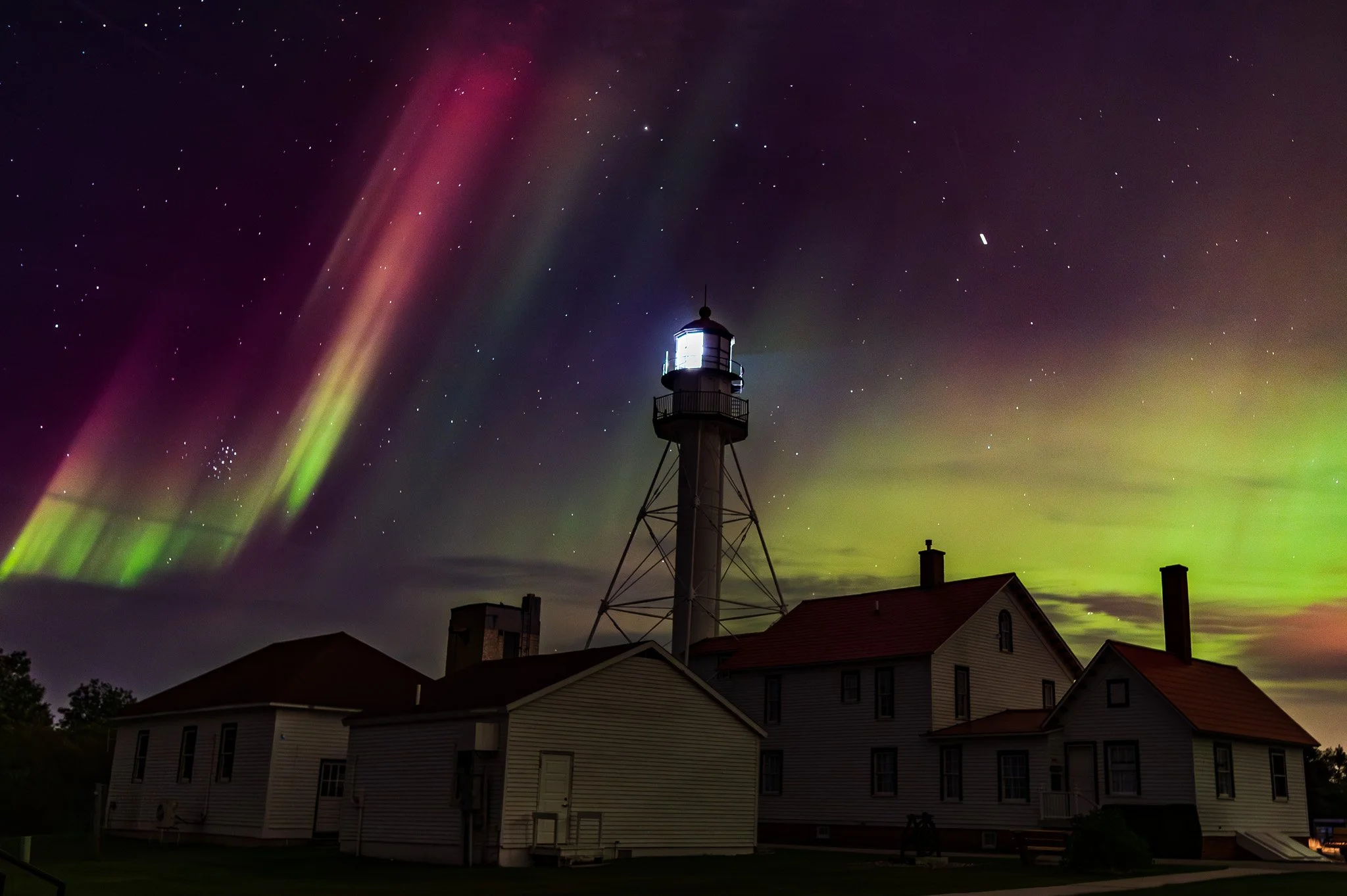 Night sky illuminated by the Northern Lights with a lighthouse and residential buildings in the foreground.