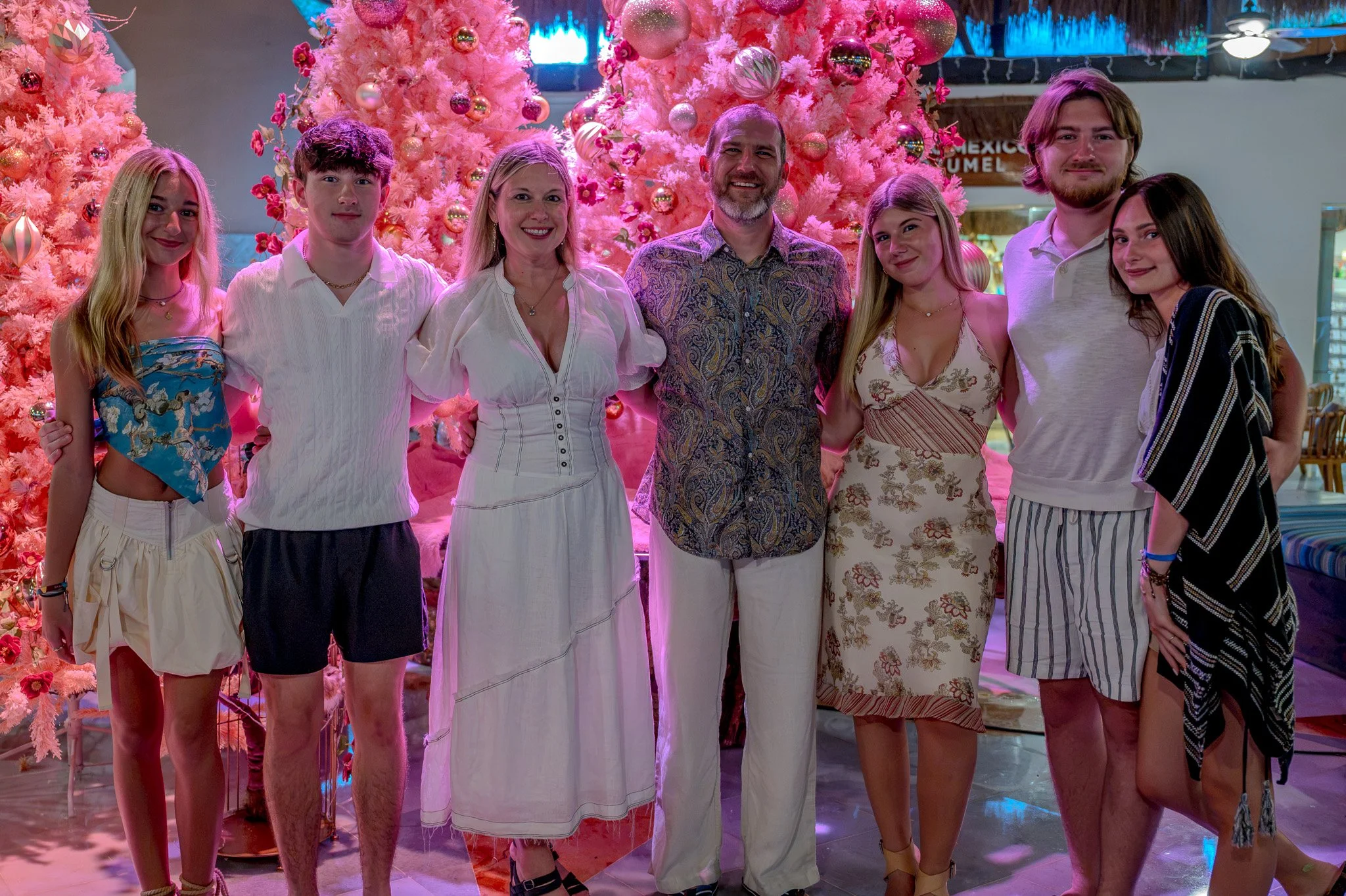 Group of eight people standing in front of a pink Christmas tree with ornaments, smiling at the camera.