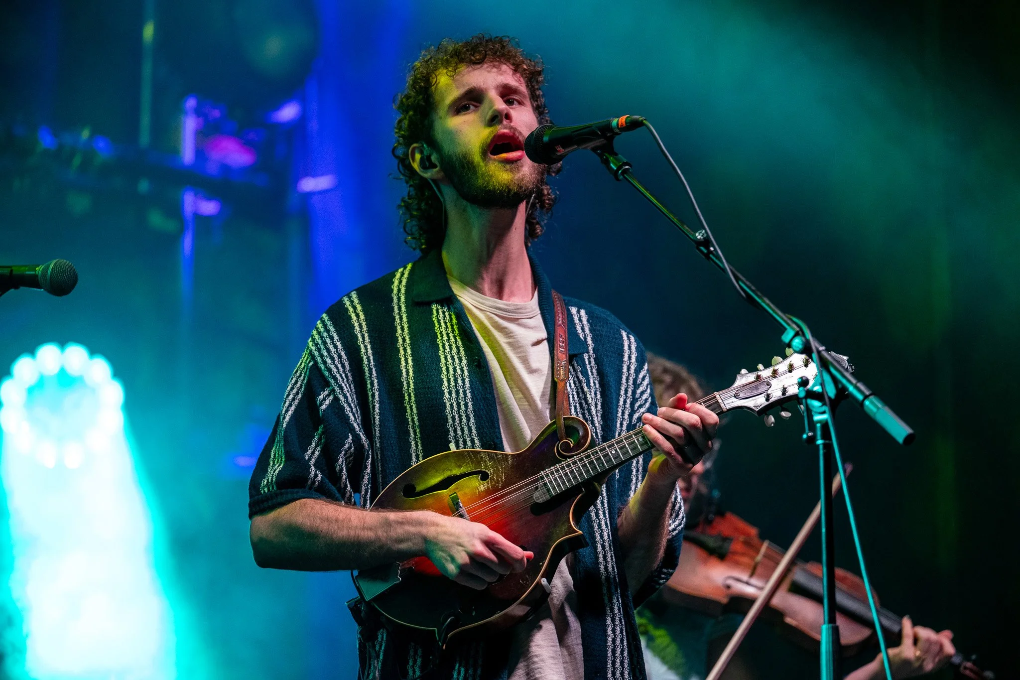 A man with curly hair and a beard singing into a microphone while playing a guitar on stage with colorful lighting.