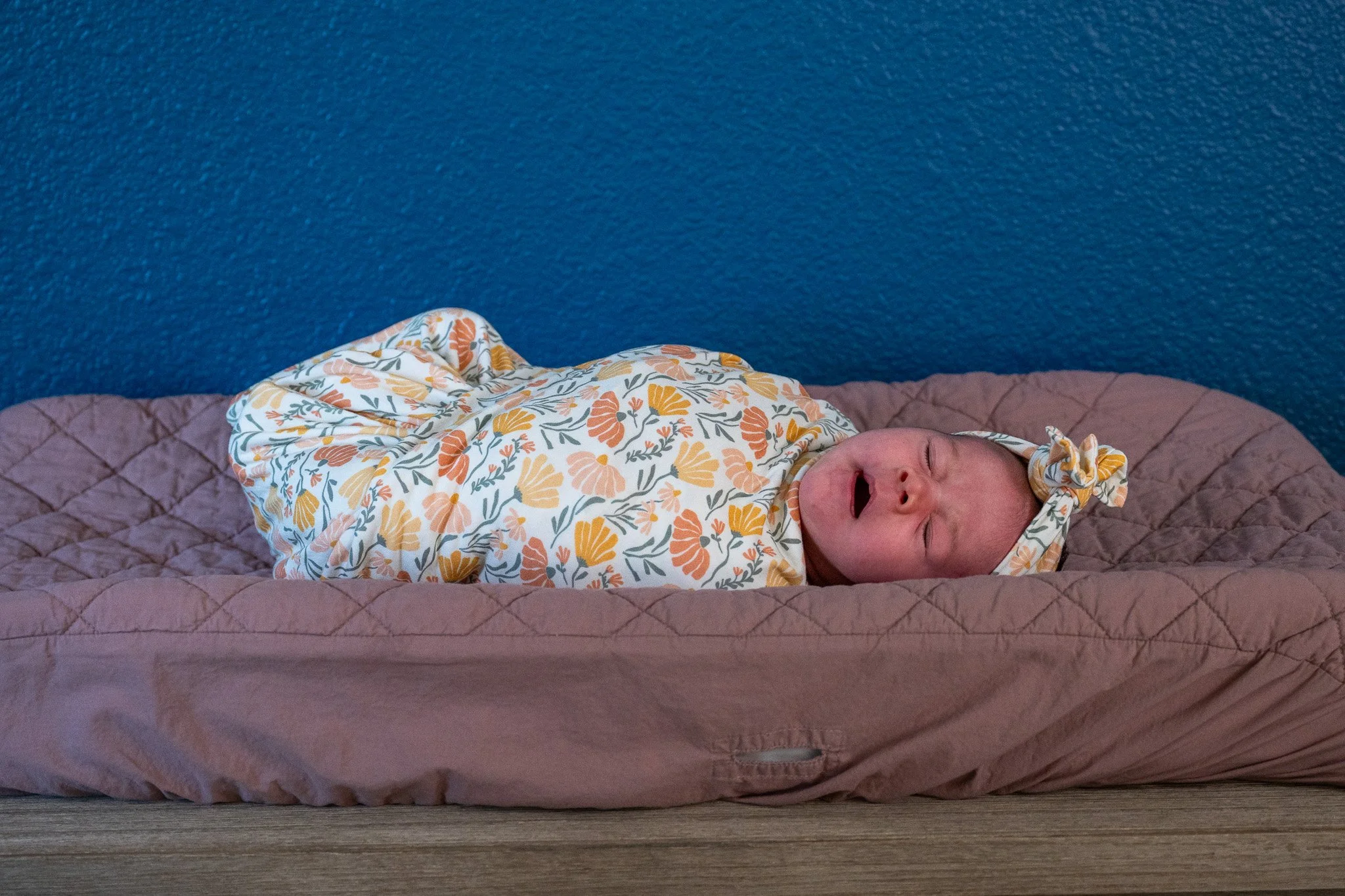 A newborn baby sleeping on a pink quilted blanket on a wooden surface, wrapped in a floral-patterned onesie with yellow, orange, and white flowers.