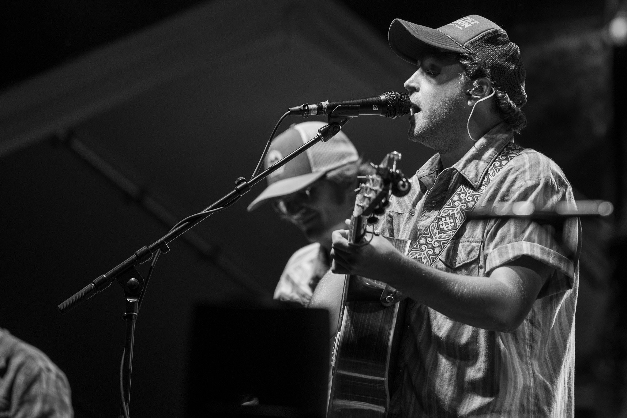 A black-and-white photo of a young man singing into a microphone while playing an acoustic guitar. He is wearing a cap and a patterned shirt, with another person partially visible in the background.