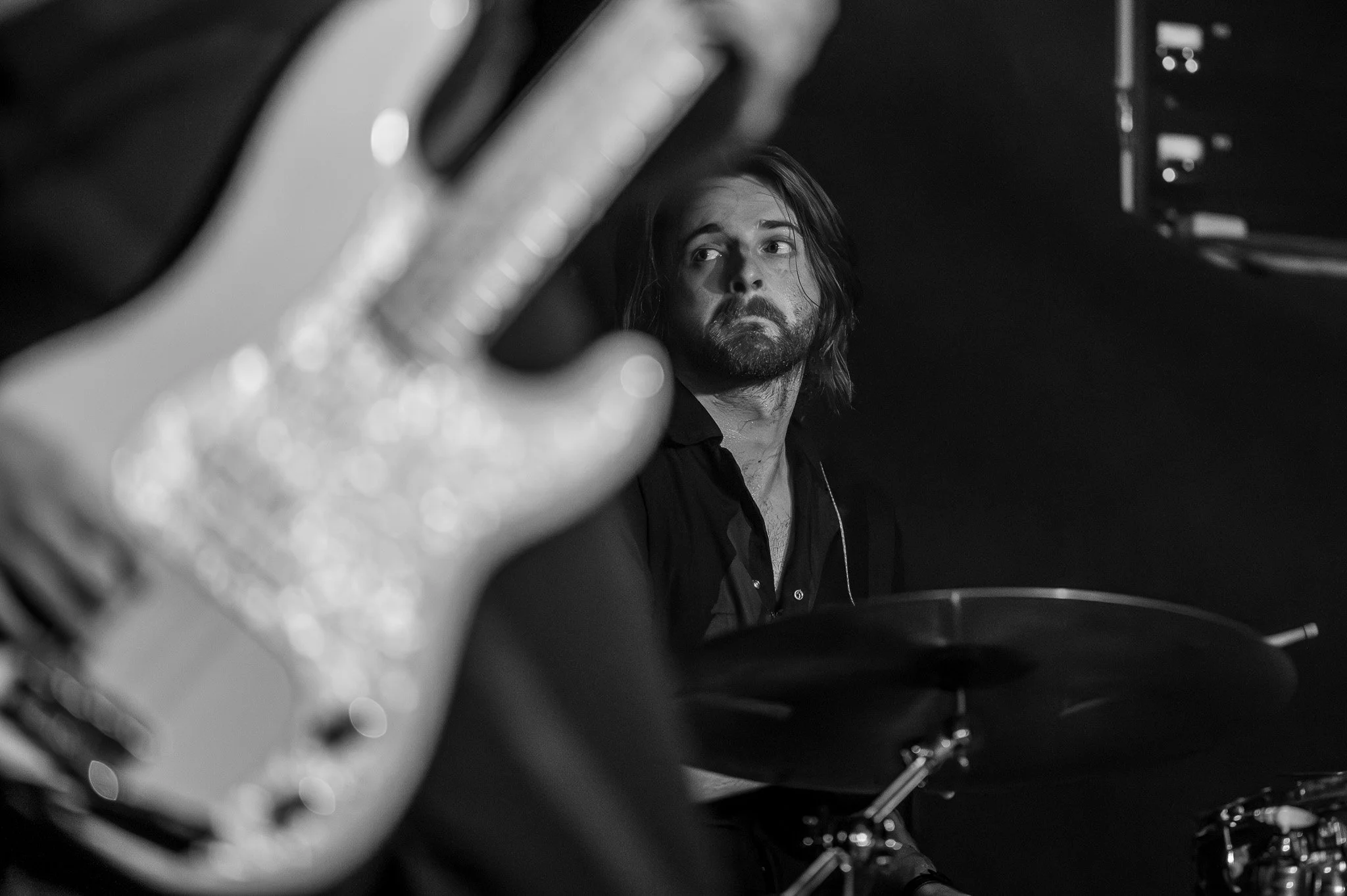 Black and white photo of a man with long hair and beard sitting at a piano, viewed through a blurred hand with rings in the foreground.