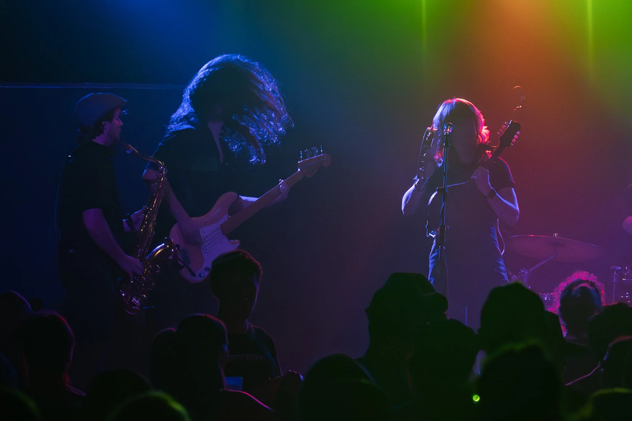 Three musicians performing on stage with colorful lights. One is playing saxophone, another guitar, and the third is singing into a microphone, with an audience in the foreground.