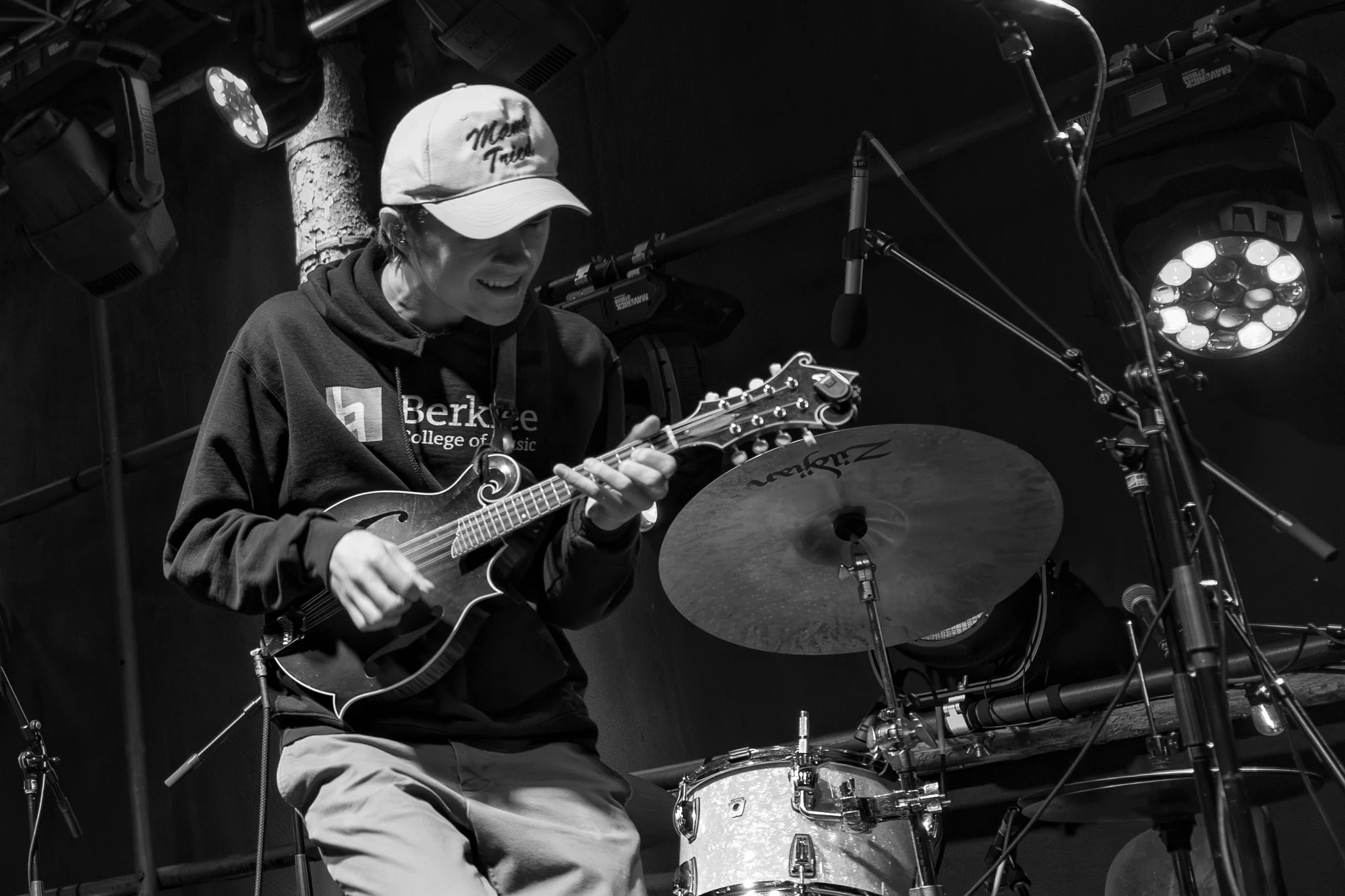 A person wearing a baseball cap and hoodie playing an acoustic guitar and sitting behind a drum set in a music studio.