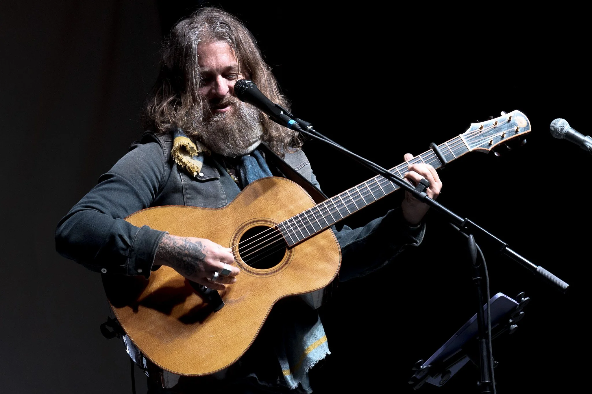 A man with long hair and a beard playing an acoustic guitar on stage, singing into a microphone, with another microphone in front of him.