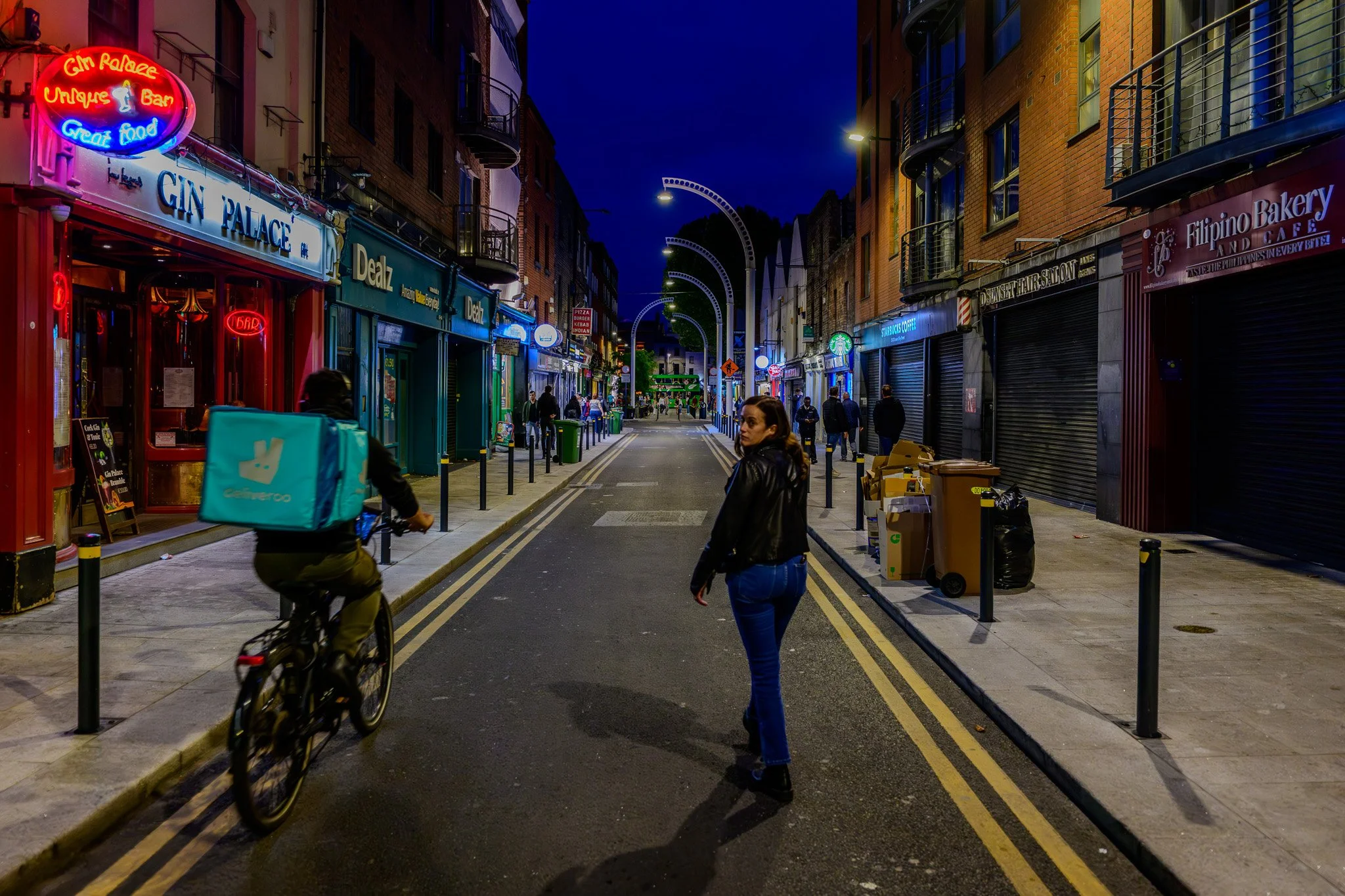 Dublin nighttime street scene with storefronts, neon signs, and pedestrians. A person is riding a bicycle, and a woman is walking. Shops include a bakery, coffee shop, and bar. 