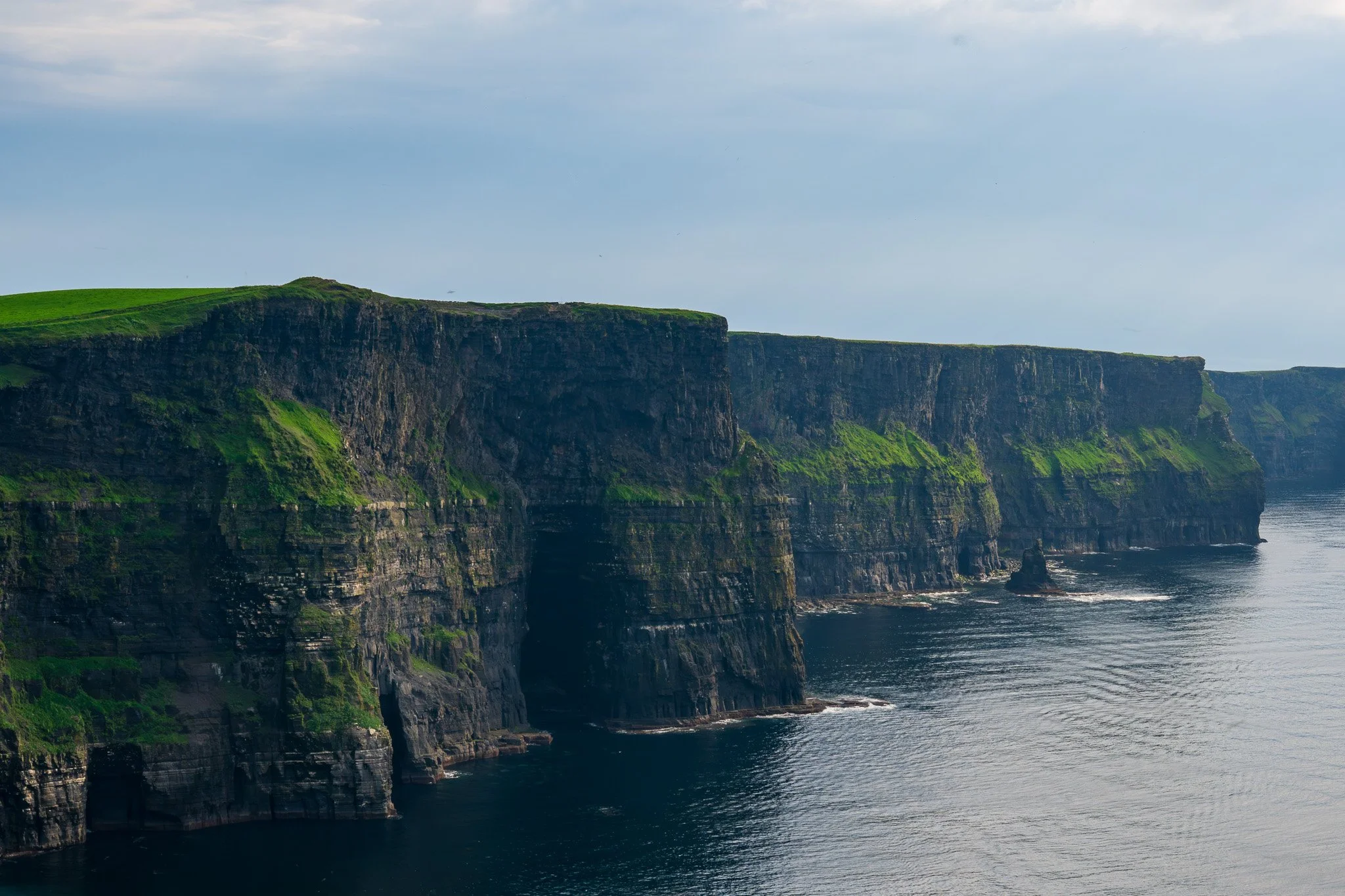 Cliffs along the coast with green vegetation on top, downward slope to rocky shoreline, ocean waves at the base, under a cloudy sky.