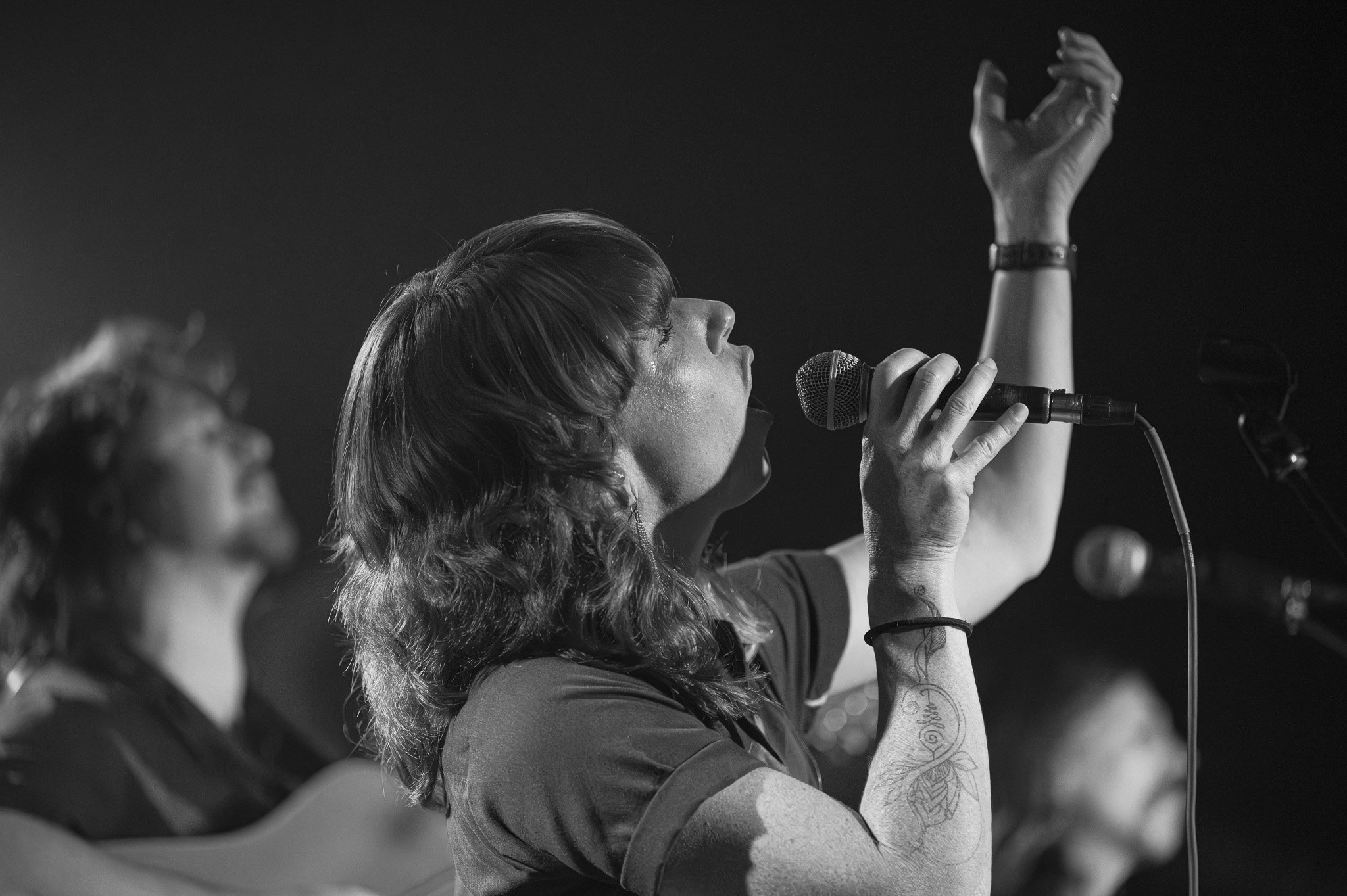 A woman singing into a microphone at a concert, with her eyes closed and her right arm raised. She has tattoos on her left arm and is wearing a watch. Other singers are visible in the background.