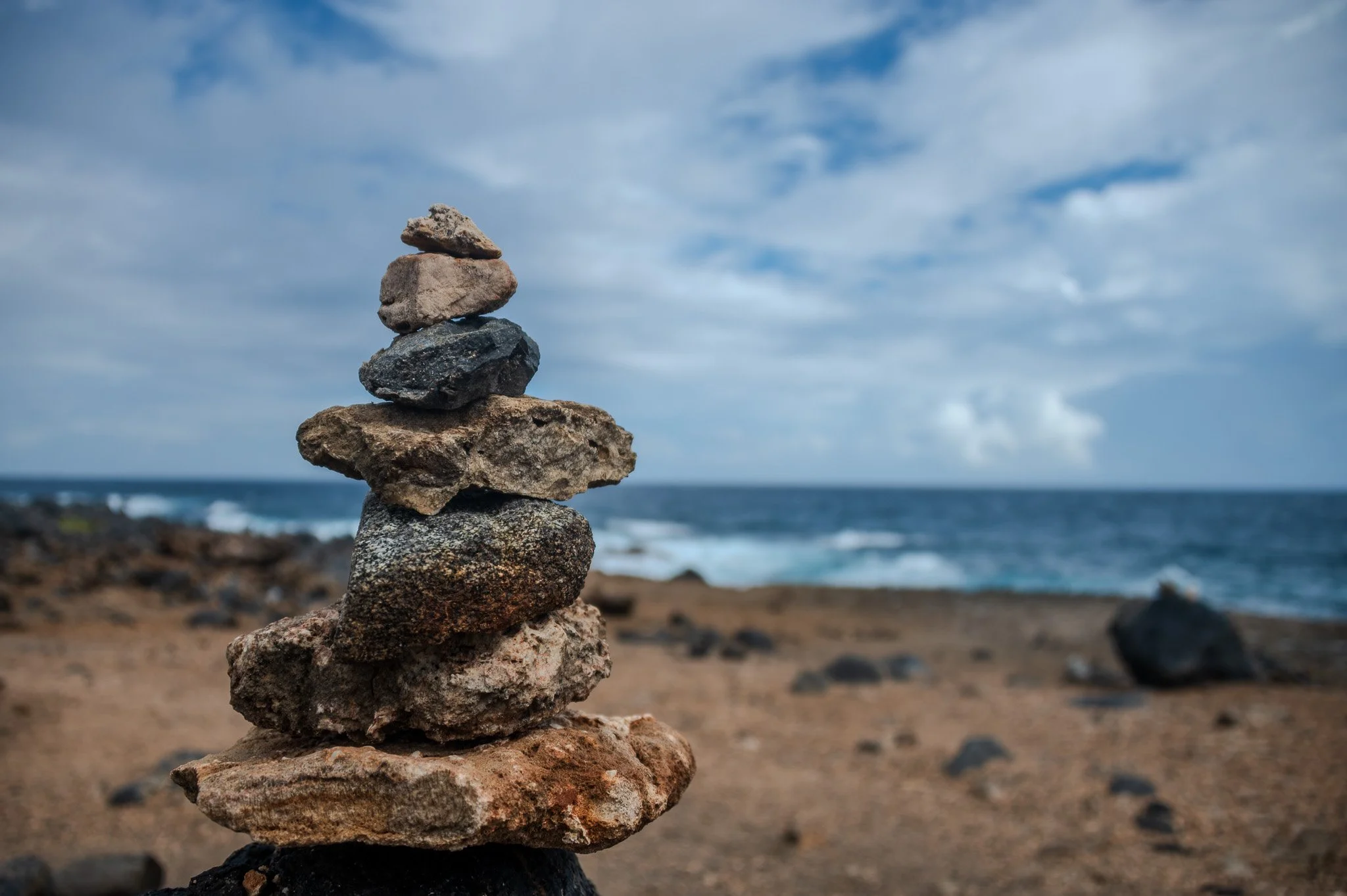A stack of seven balanced rocks at a beach with ocean waves and partly cloudy sky in the background.
