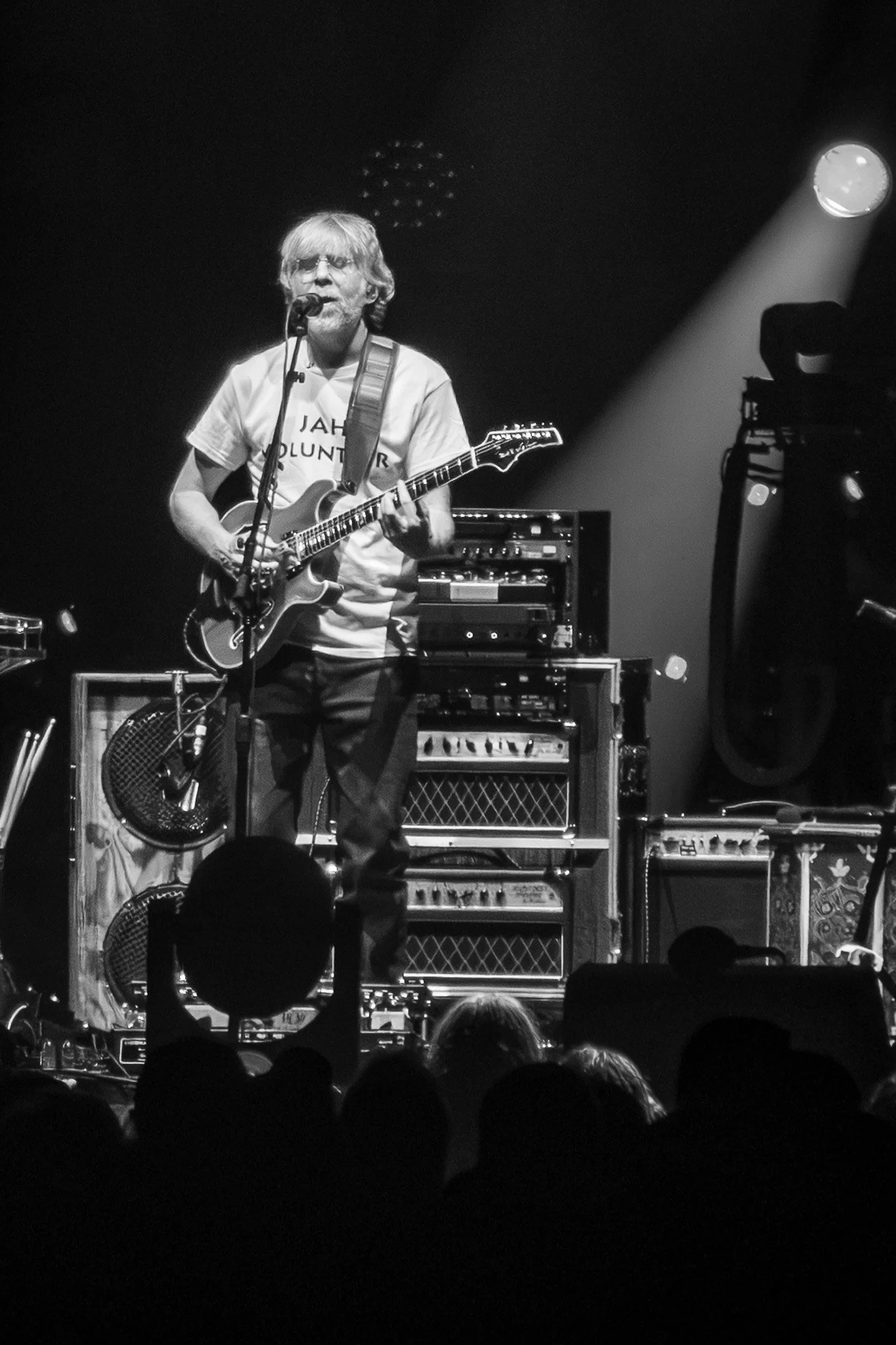 A black-and-white photo of a male musician singing into a microphone while playing an electric guitar during a live concert. The scene includes stage equipment, an amplifier, and the silhouette of an audience with some raised hands.